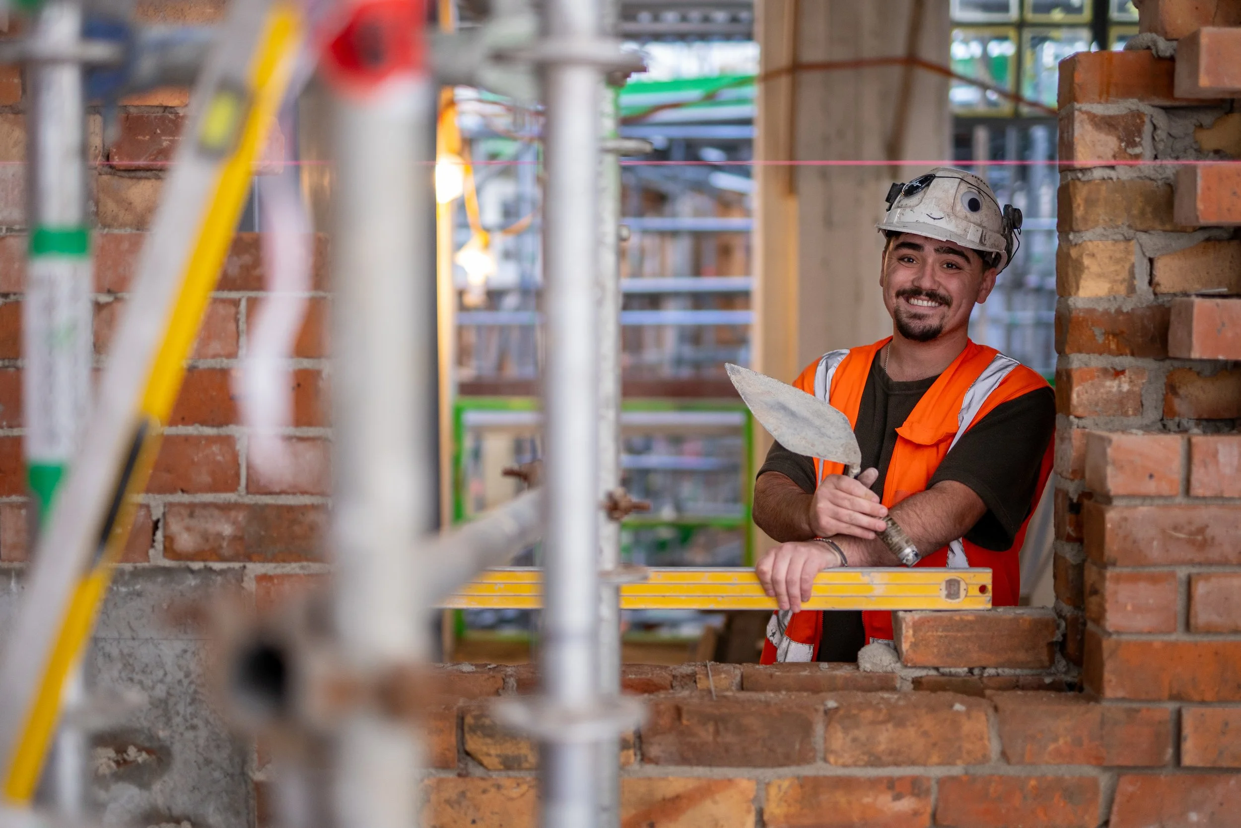 A construction worker with a beard and smiling face wearing a white safety helmet and an orange safety vest, holding a trowel, standing at a brick wall under construction, with construction scaffolding and building materials visible in the background