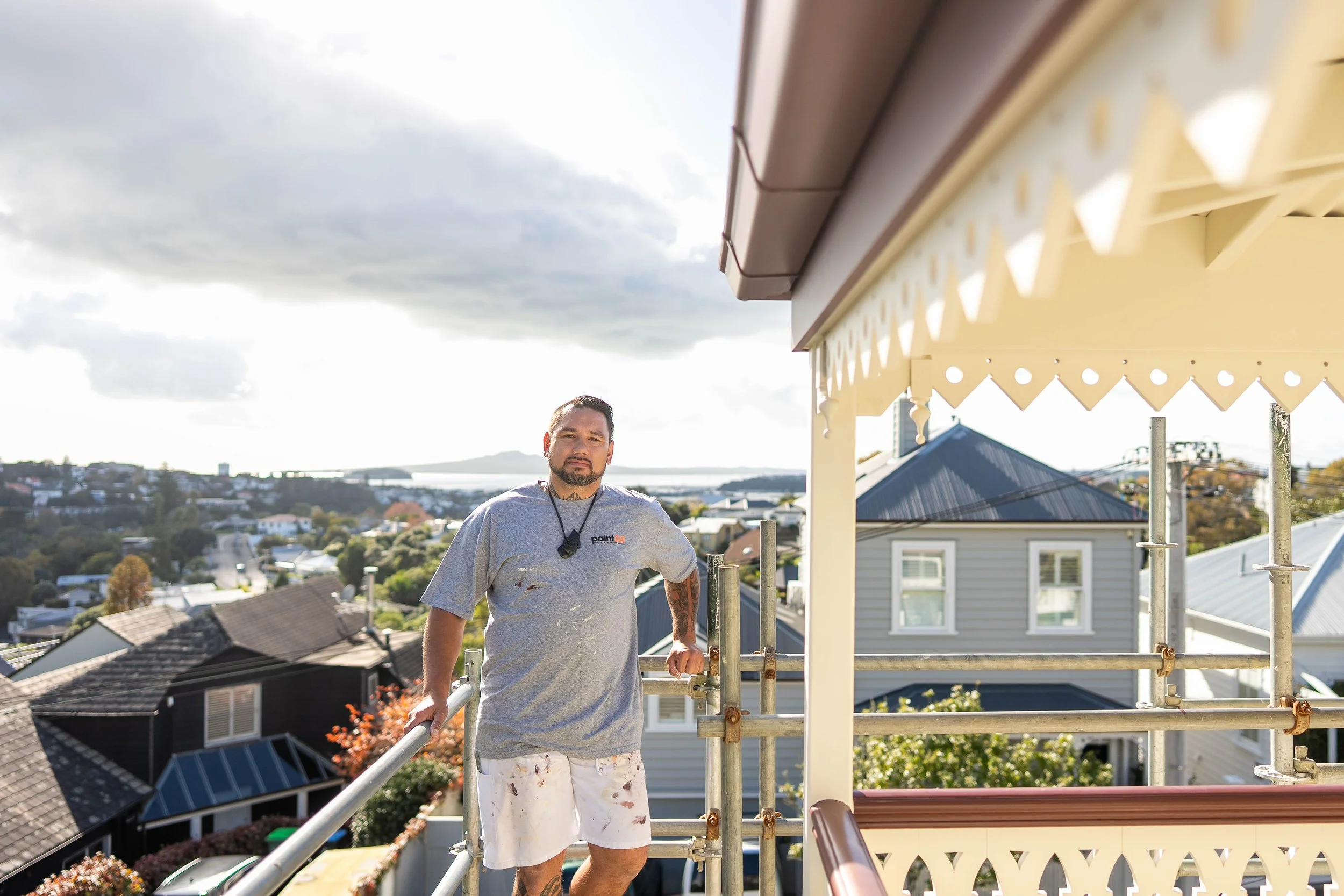Man standing on a scaffolding on a balcony during daytime, with houses and a distant view of water in the background.