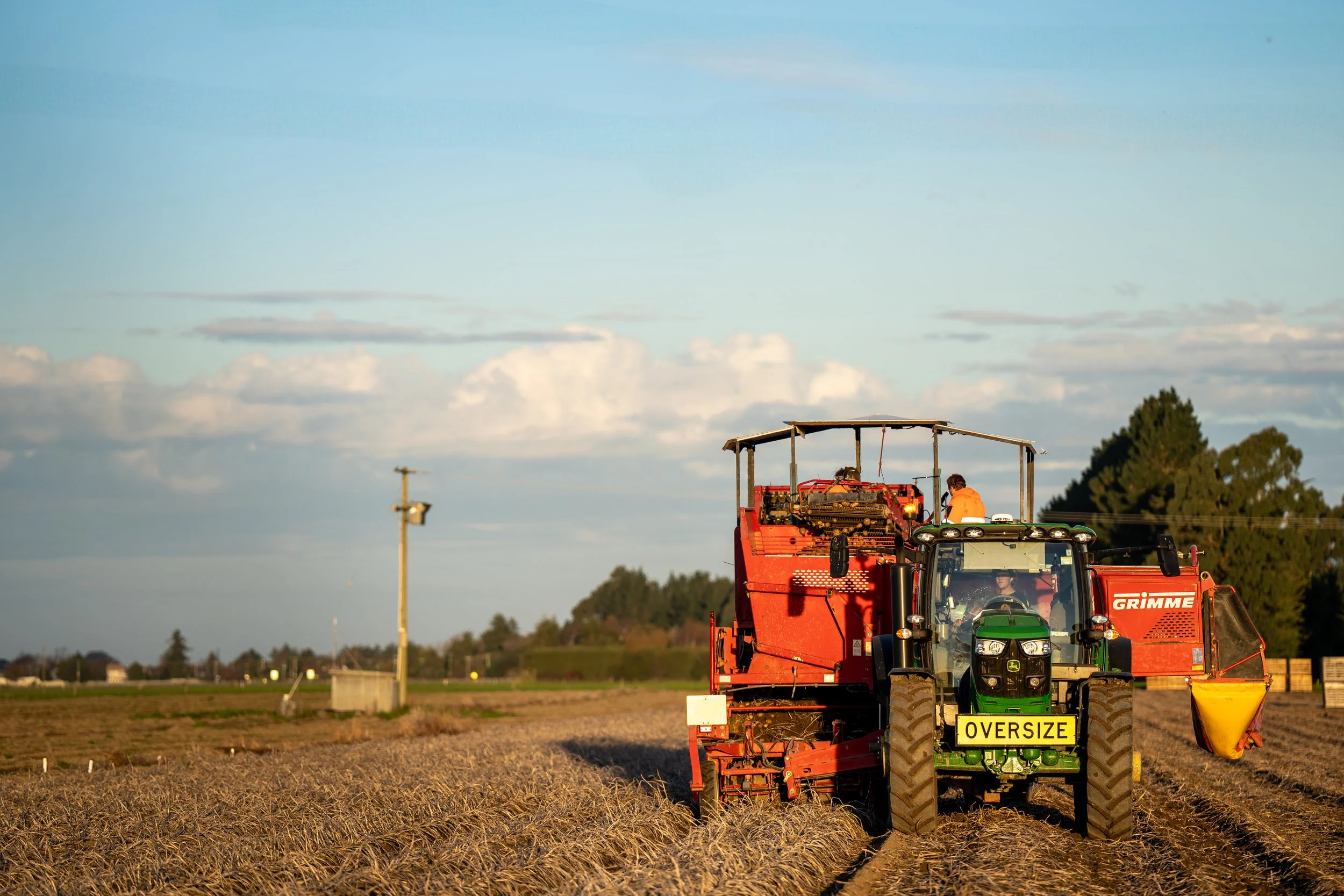 A green tractor with an oversized sign is driving through a field, attached to red farming equipment, with workers standing on top. The field appears to be harvested or in the process of harvesting, under a partly cloudy sky.