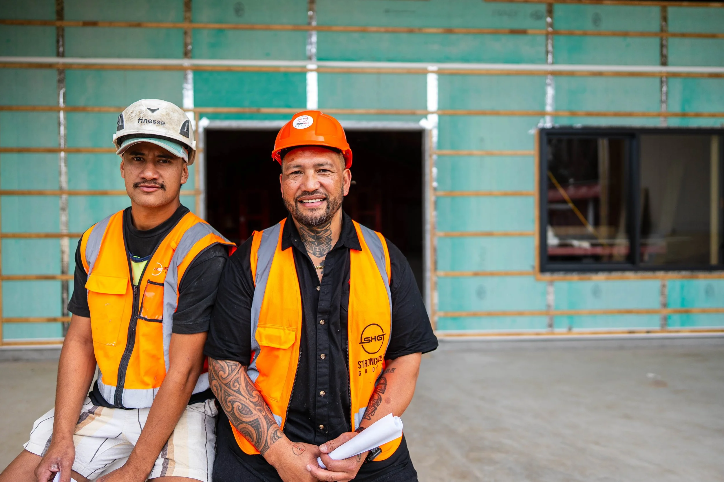 Two construction workers standing in front of a building under construction, wearing safety vests and helmets.