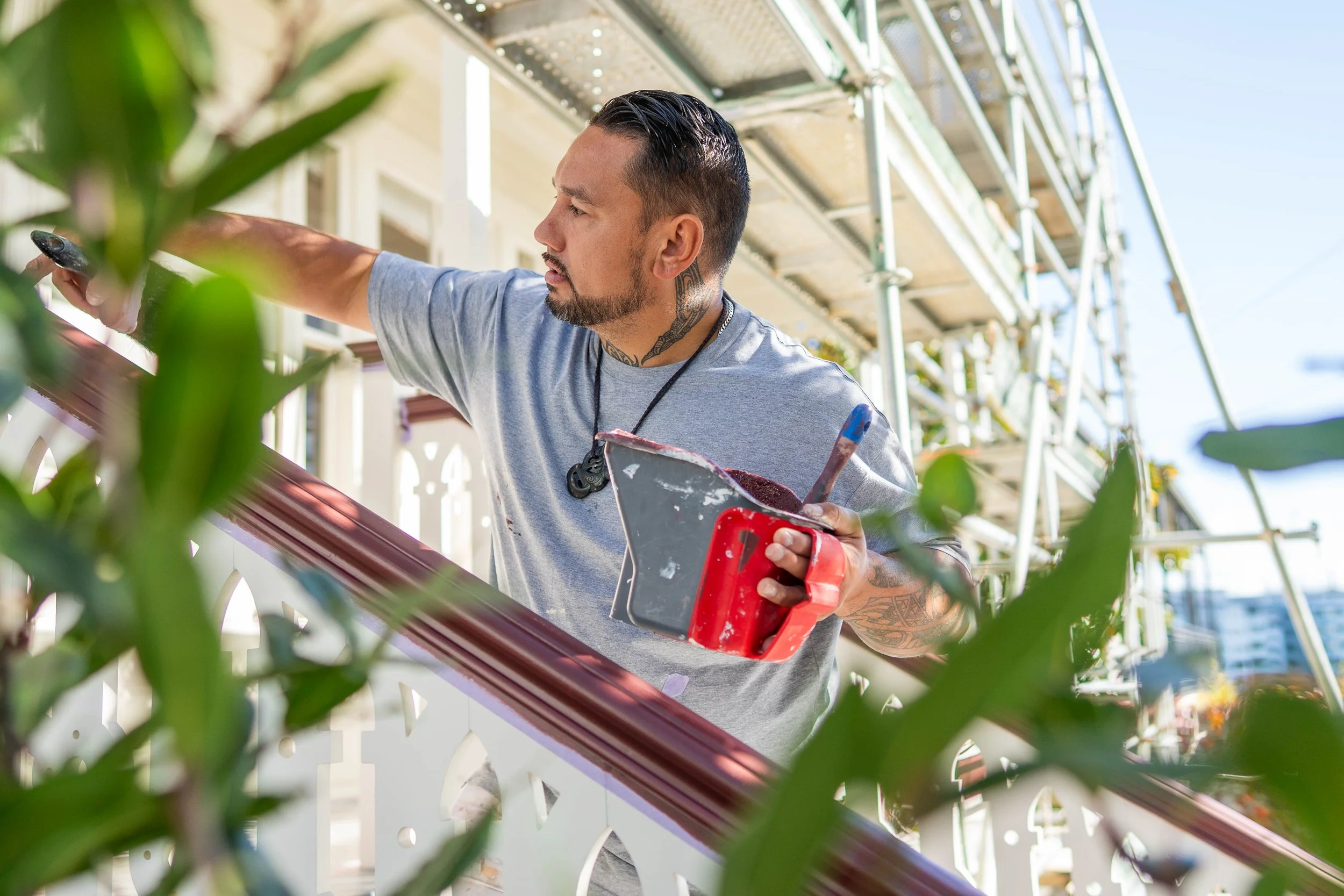 A man with tattoos on his neck and arm, wearing a gray t-shirt, is painting a decorative wooden railing on a porch or balcony. He holds a paintbrush in one hand and a red container of paint in the other, with a scaffolding structure and a clear sunny