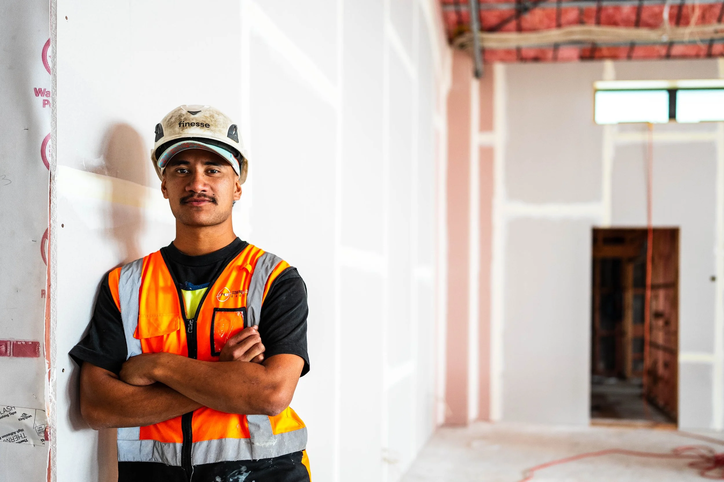 A construction worker wearing a safety helmet and orange safety vest stands with arms crossed inside a building under construction.