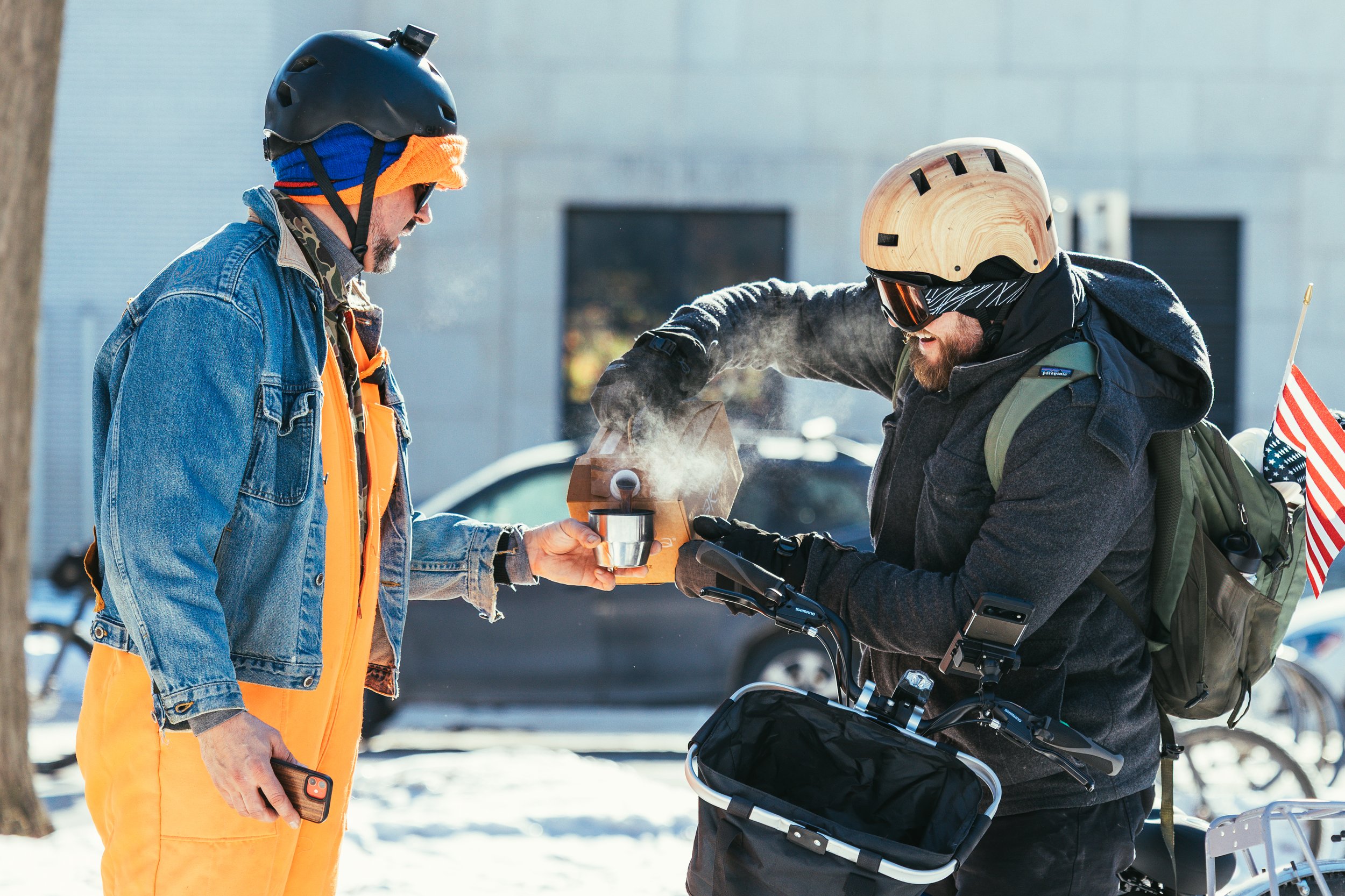 Two men attending the Alex Pretti memorial bike ride, one man pouring a steaming cup of coffee for the other, both dressed warmly, with the Minneapolis Institute of Art in the background.
