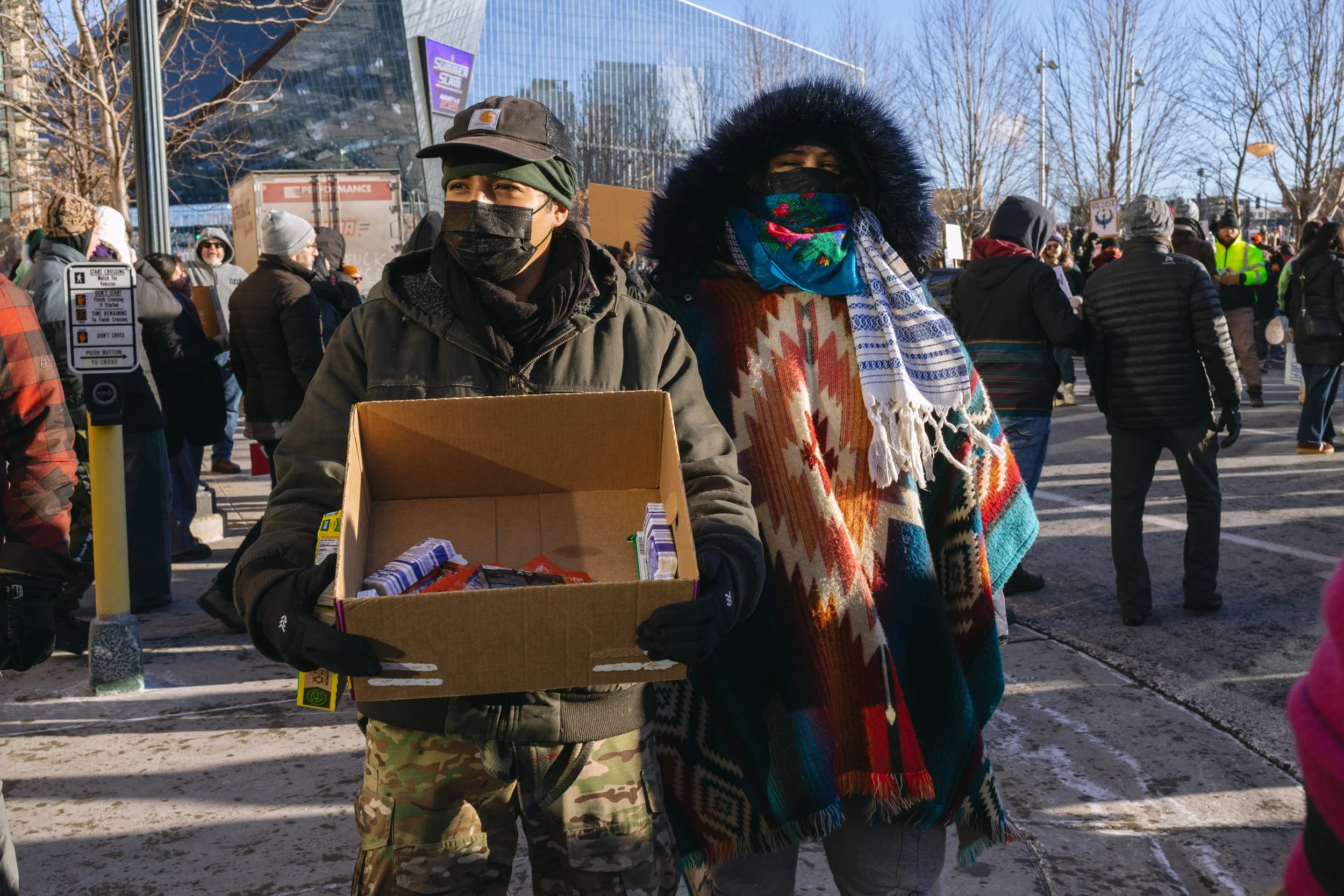 Two people wearing masks and winter clothing, standing outside at the ICE protest in Minneapolis, with many other people and US Bank Stadium in the background.