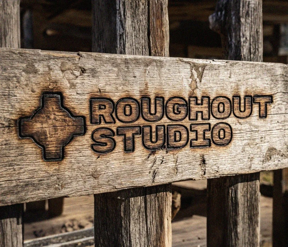 Roughout Studio, A weathered wooden sign with the logo of Texas shaped like a cross and the text "ROUGHOUT STUDIO" engraved on it.