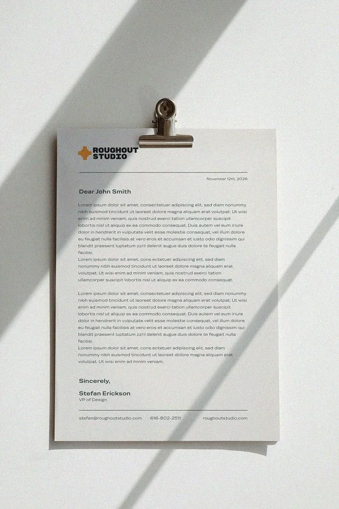 Roughout Studio, A letter on white paper clipped to a wall, with shadows cast across it. The letterhead says "ROUGHOUT STUDIO" with a small orange cross icon. The letter is addressed to John Smith and is signed by Stefan Erickson, VP of Design.