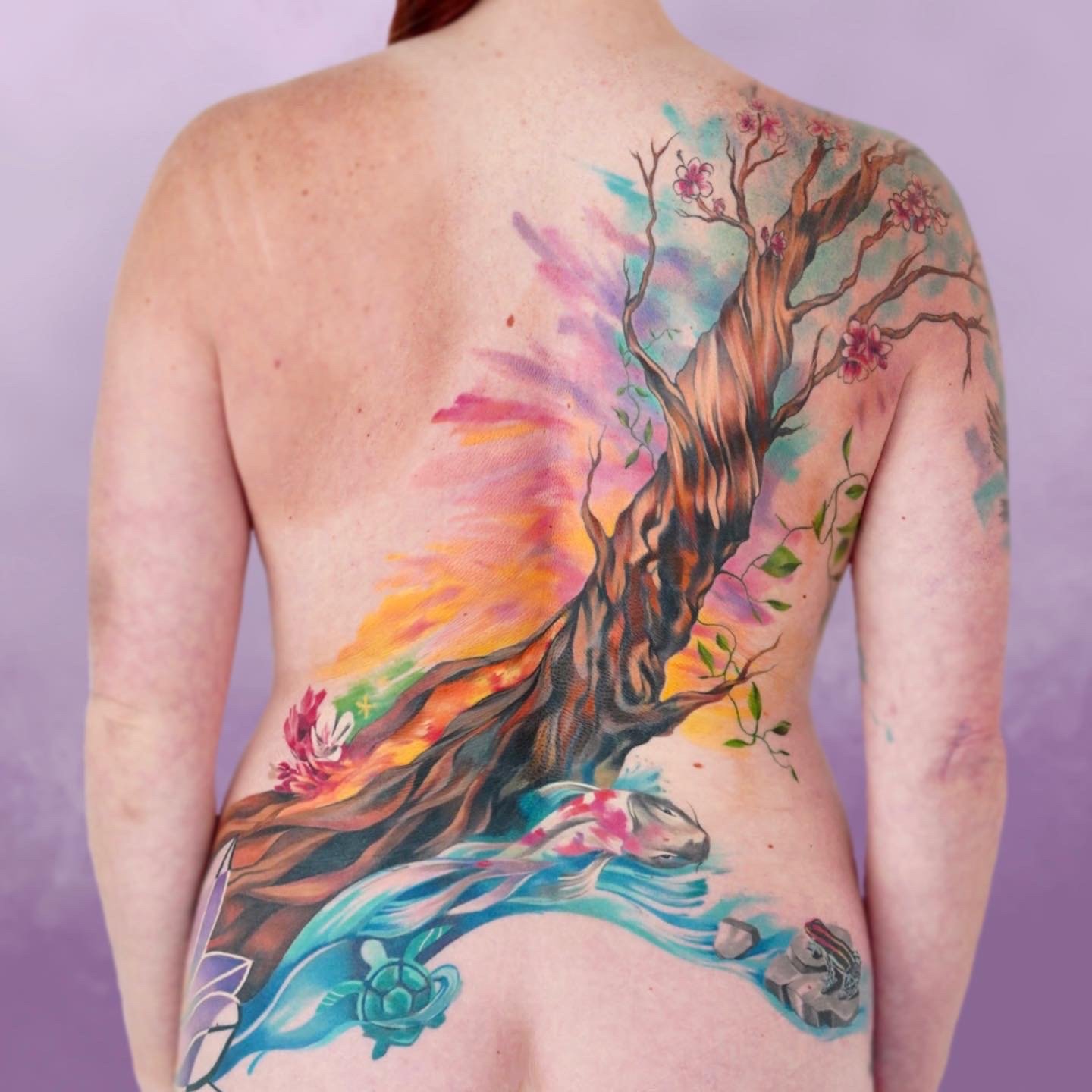 Colorful tattoo of a large tree with pink blossoms, flowing water at its base, and a turtle on a rock on a person's back and side.