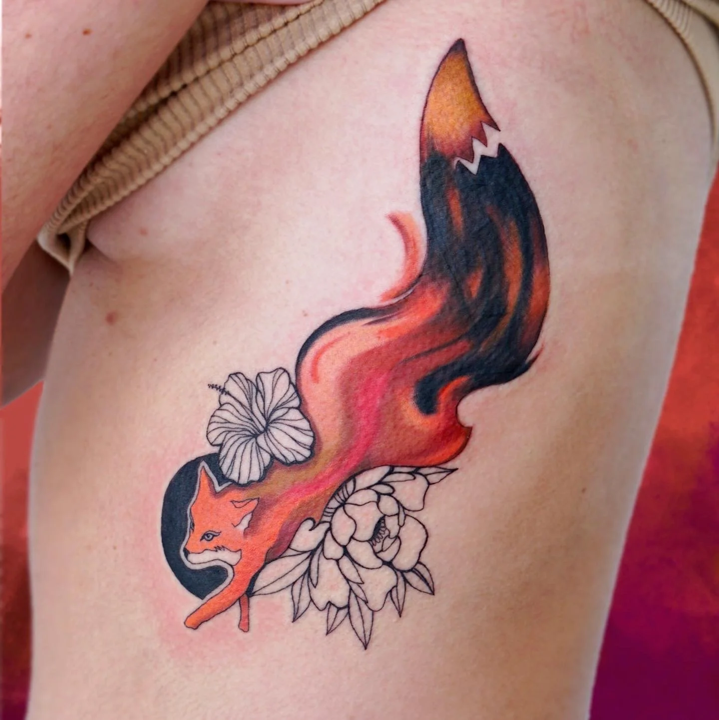Tattoo of a fox's head and tail in watercolor style, surrounded by flowers, on person's ribcage.