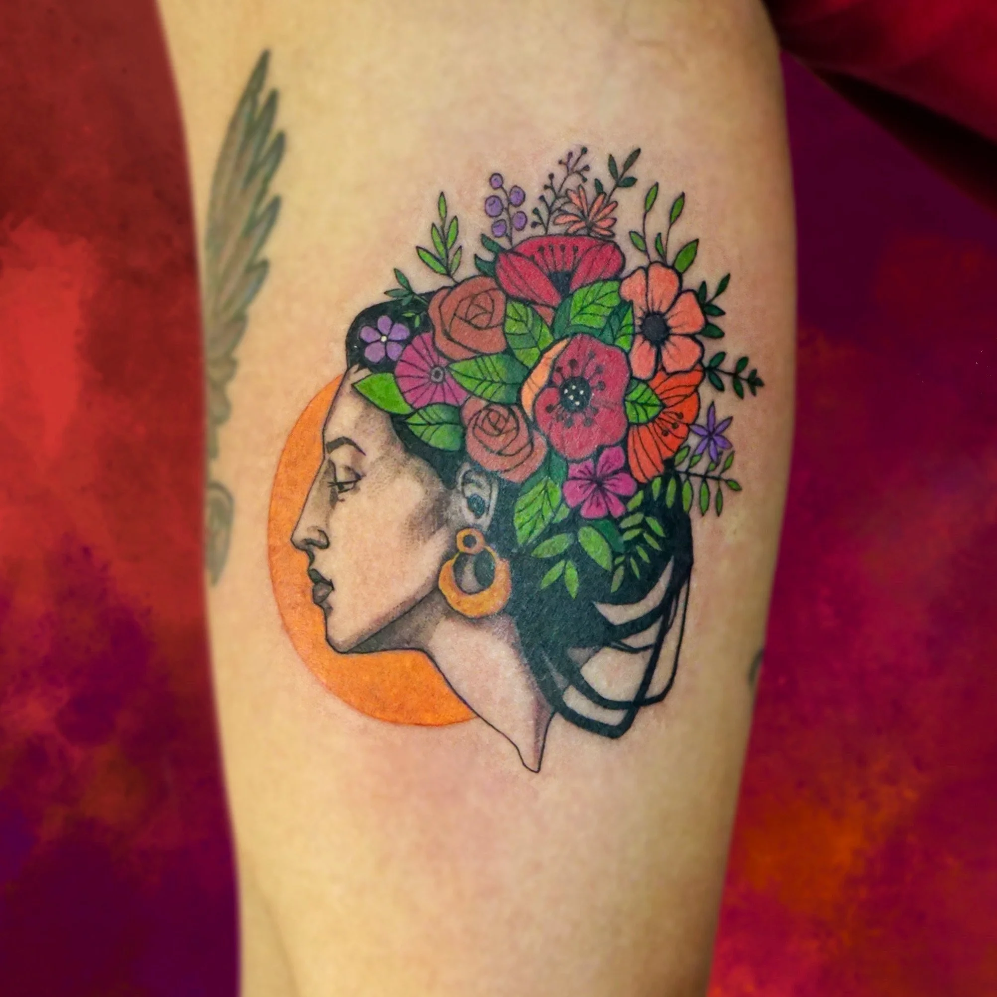 Tattoo of a woman's profile with long hair, earrings, and a crown of colorful flowers, with a red-orange sun or moon in the background.
