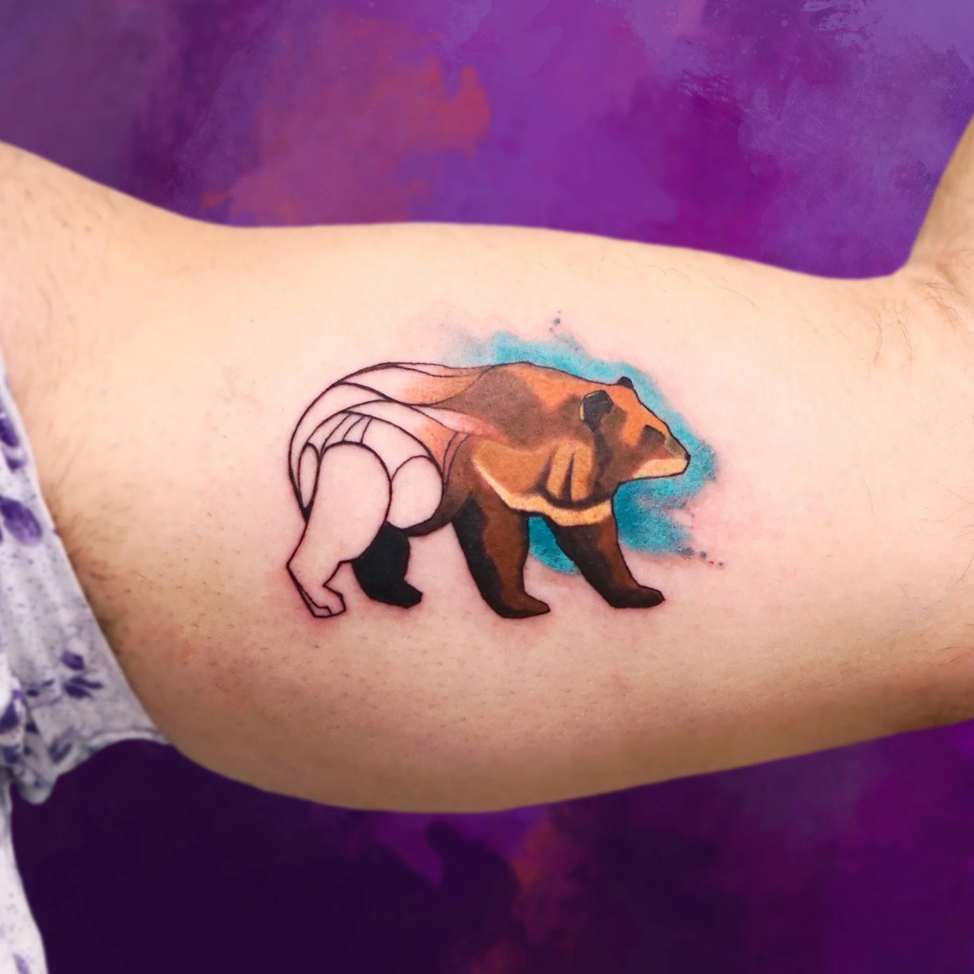 Tattoo of a bear with wings on a person's arm, with a watercolor-style purple background.