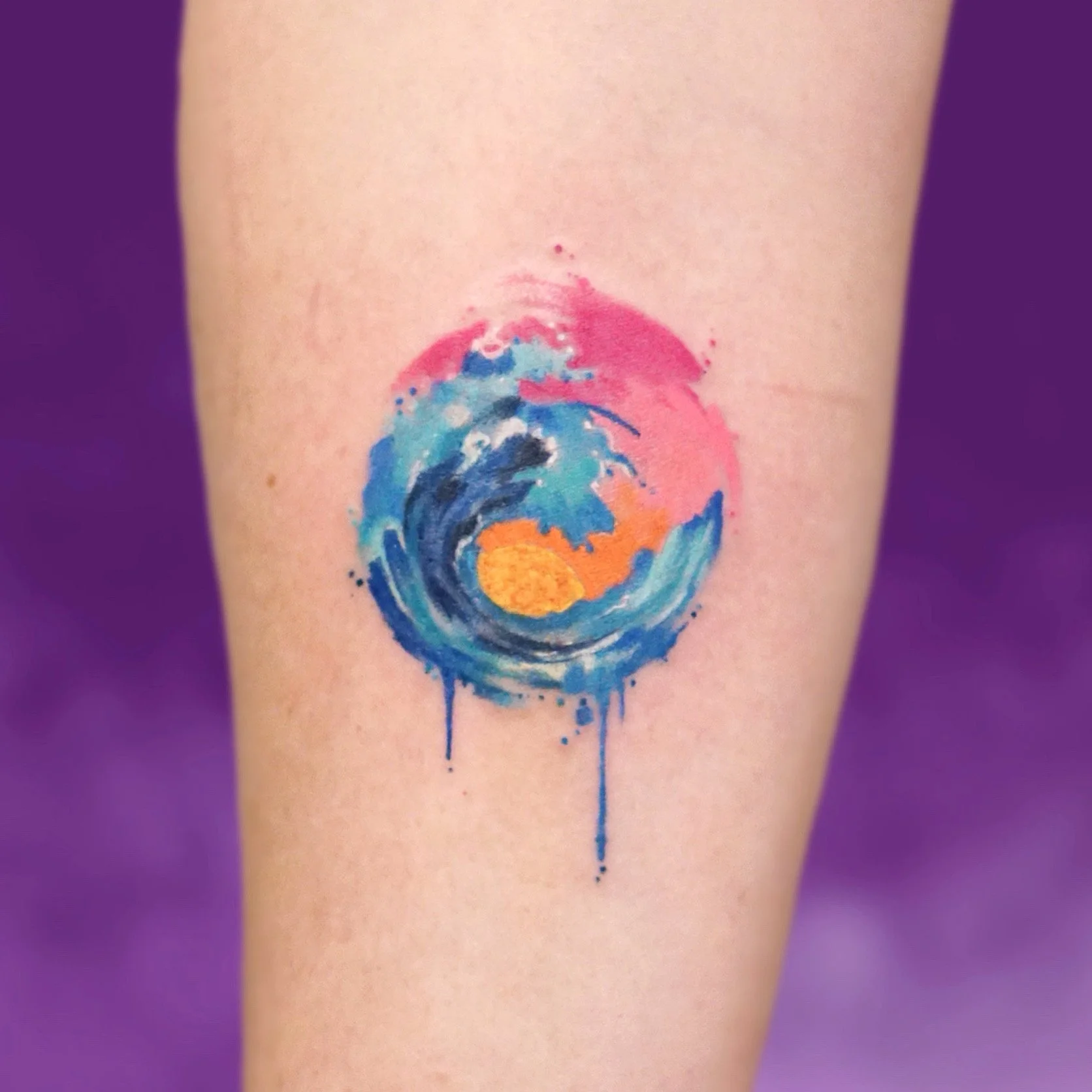 Watercolor-style tattoo of a wave around a sun, with pink and blue hues, on a person's arm.