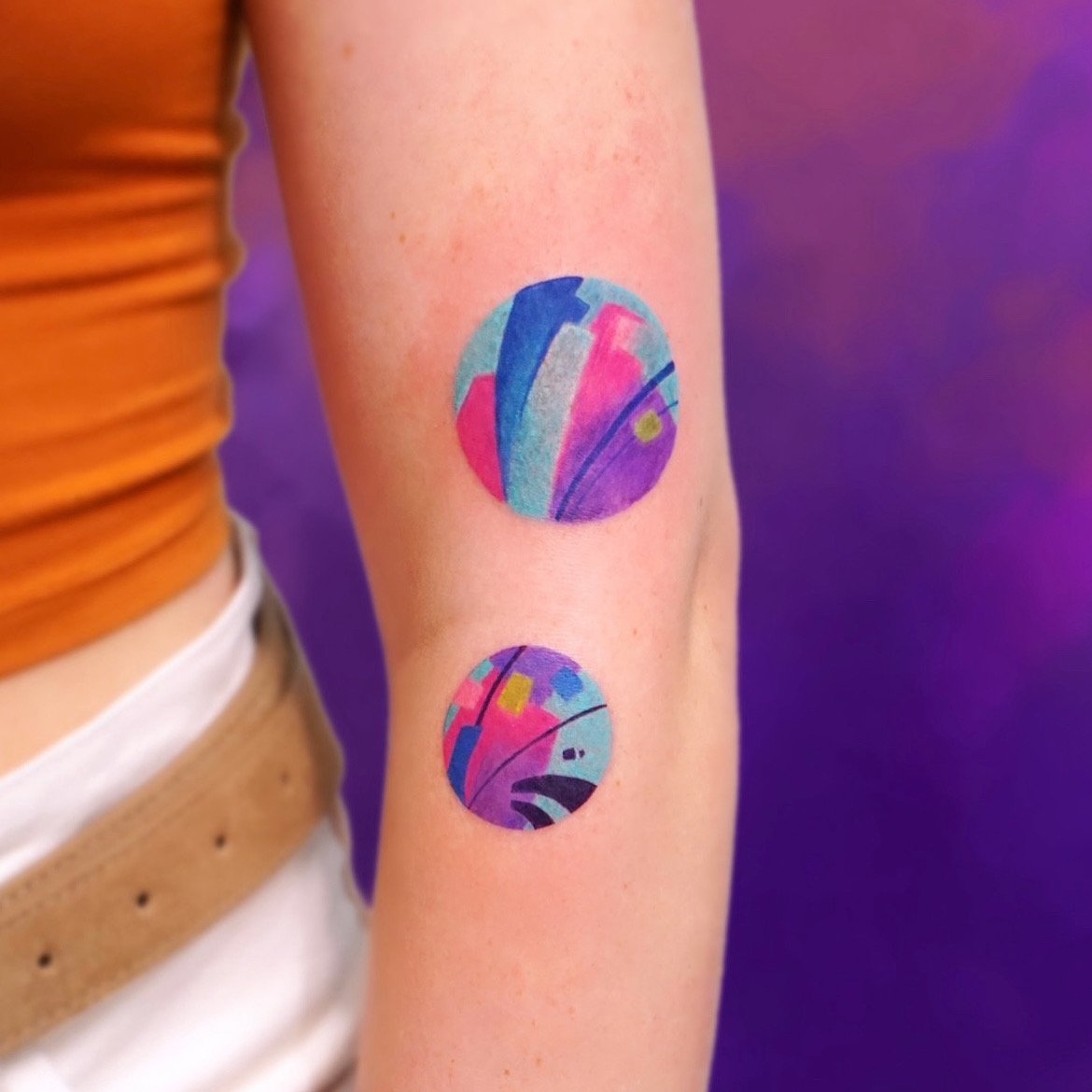 Two colorful, abstract circular tattoos on a person's arm with a purple background.