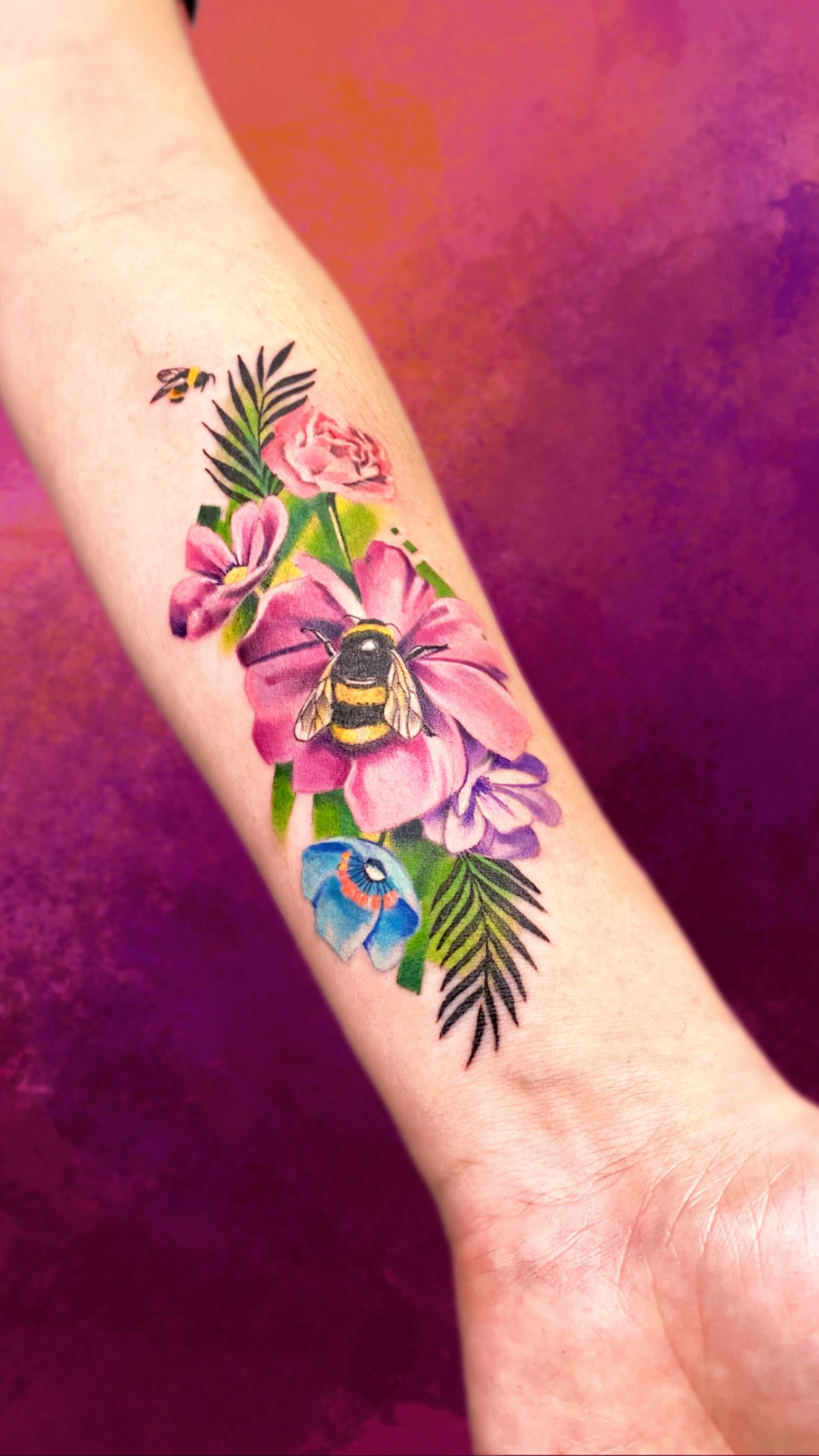 A colorful tattoo featuring pink, purple, and red flowers, green leaves, a bee, a butterfly, and a small yellow and black insect on an arm with a pink and purple background.