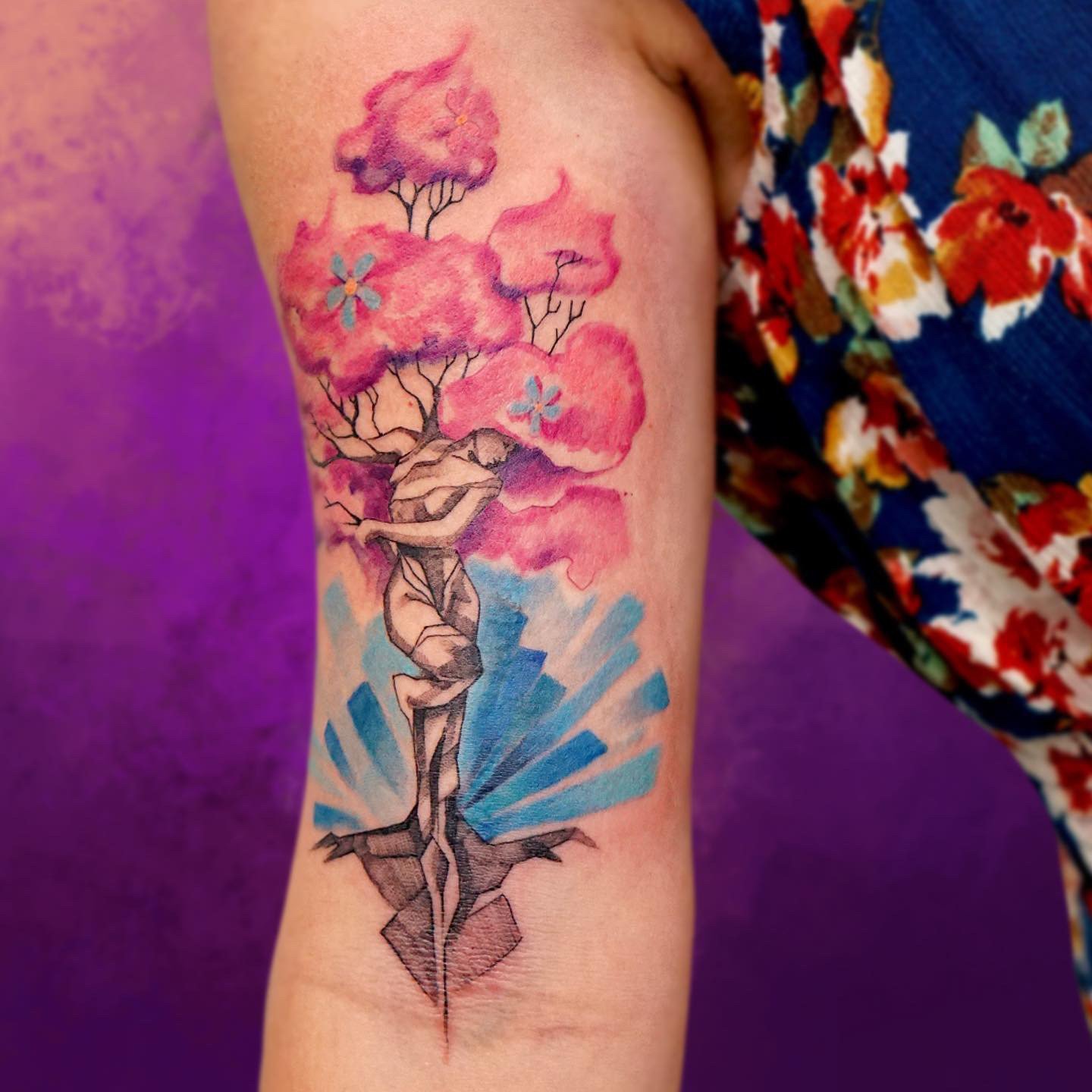 A colorful watercolor tattoo of a tree with pink, purple, and blue foliage, a woman with long hair merged with the tree trunk, and small blue flowers, on a person's upper arm with a purple and pink background.