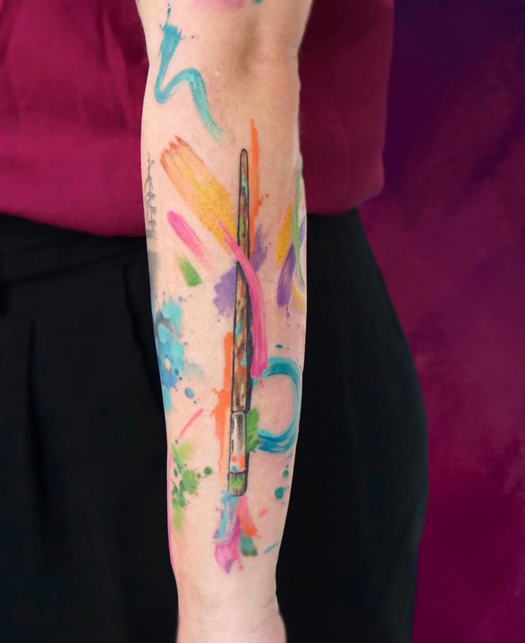 Colorful watercolor tattoo of paintbrushes and splashes on person's forearm.