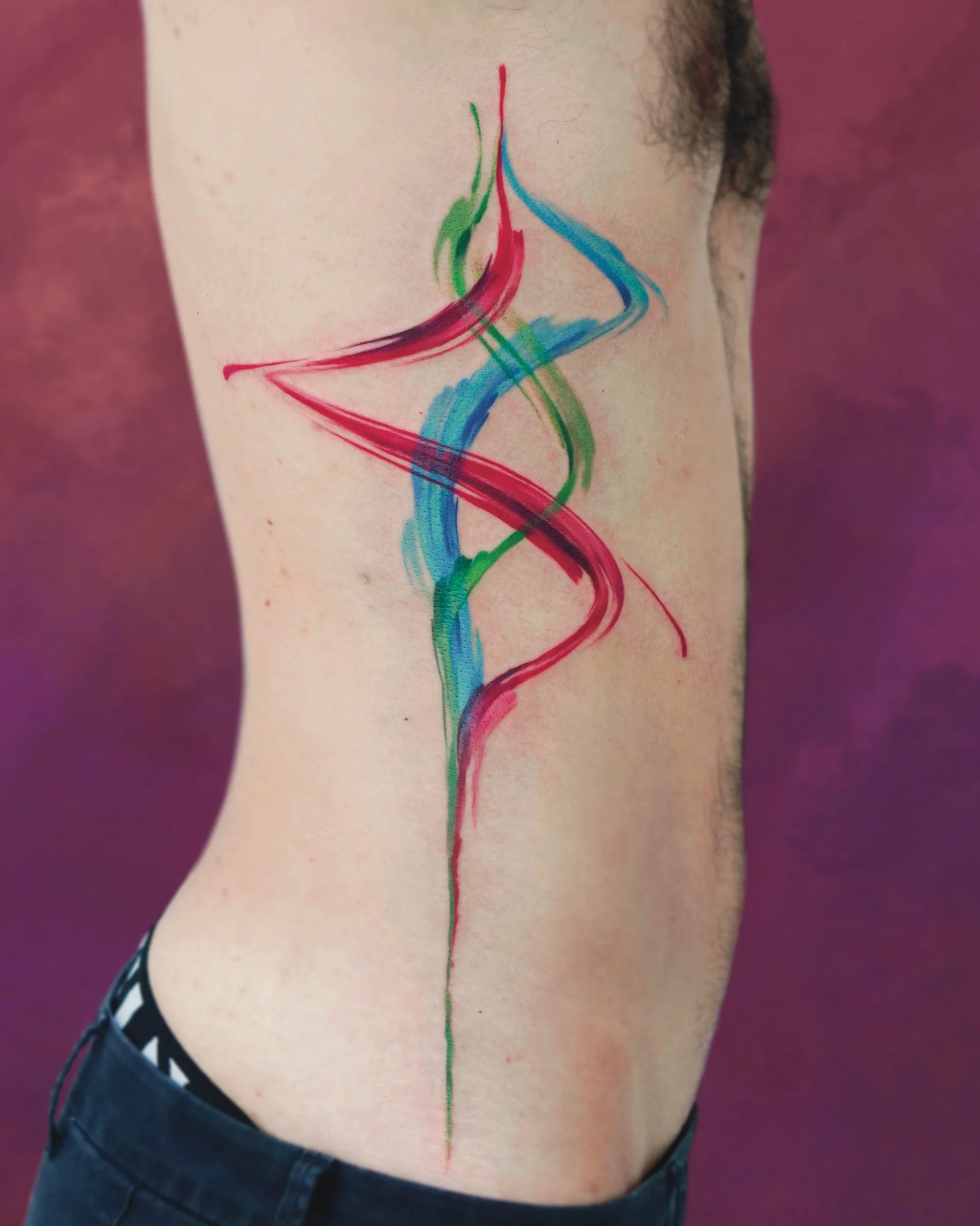 Colorful abstract tattoo on a person's side torso featuring flowing red, green, and blue lines against a blurred purple background.