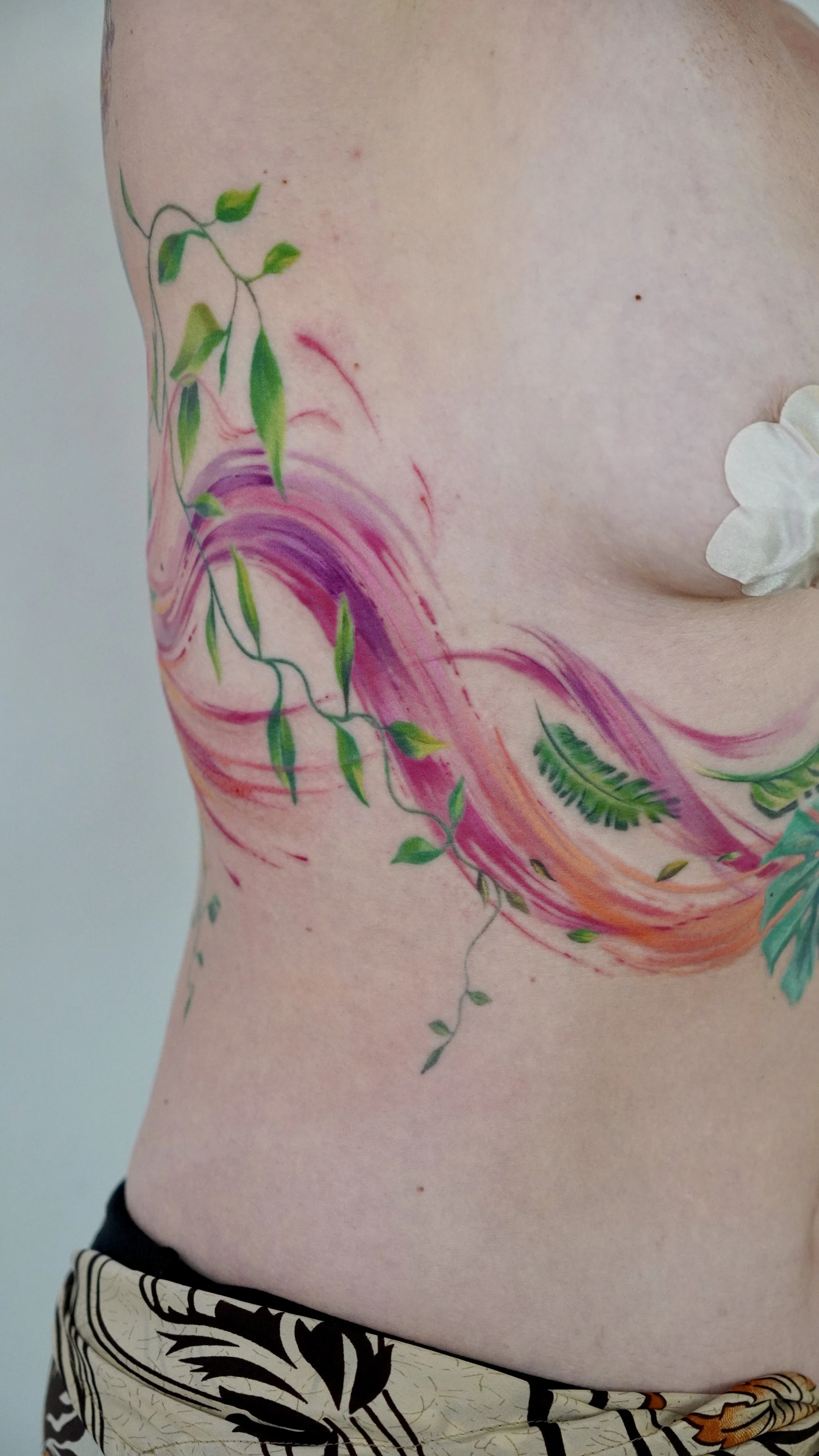 Colorful tattoo of a rainbow, green leaves, and a fern on a person's side torso.