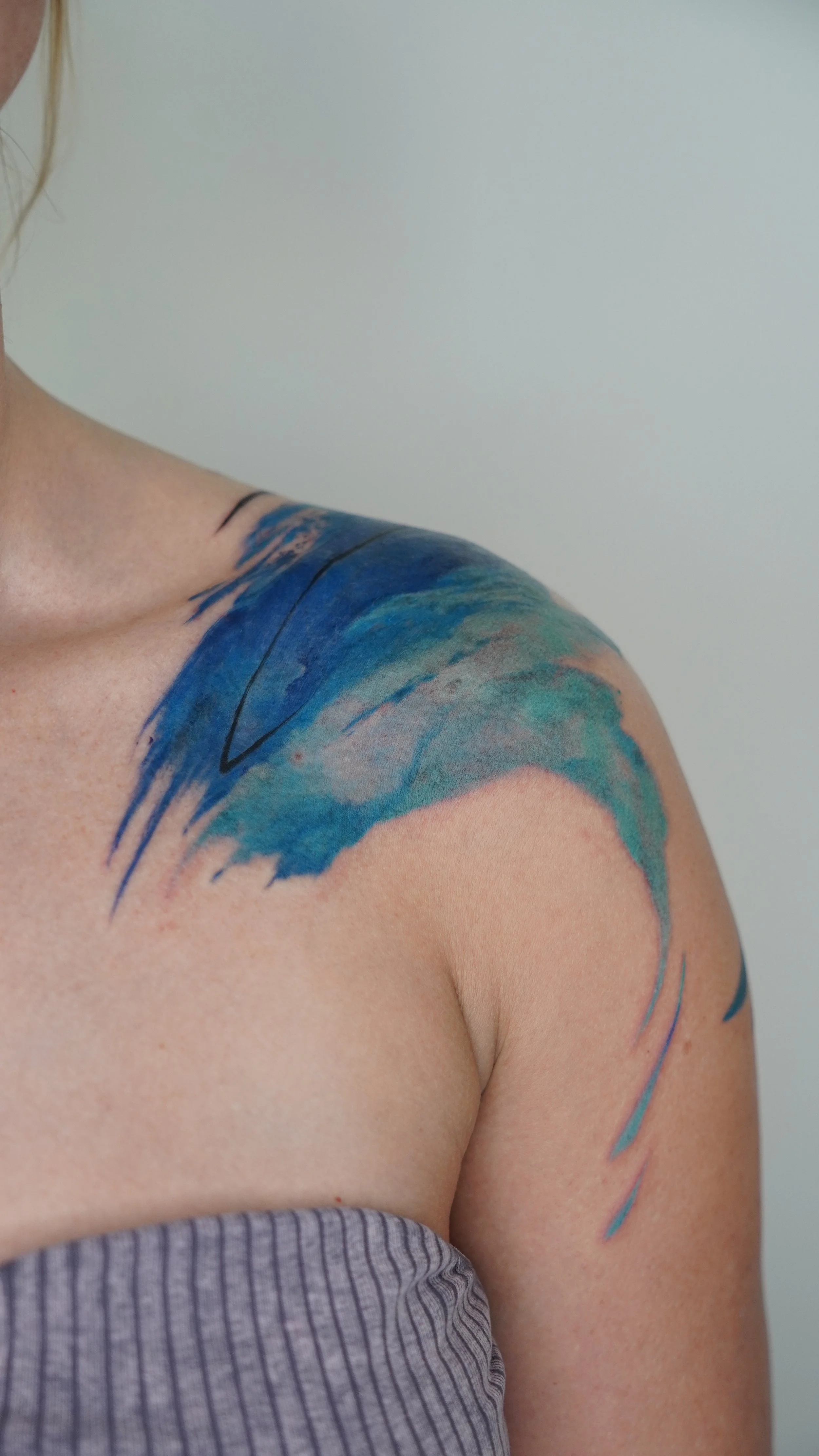 Close-up of a woman's shoulder showing a watercolor-style tattoo in shades of blue and green, with lighter areas blending into her skin, and part of a striped grey top.