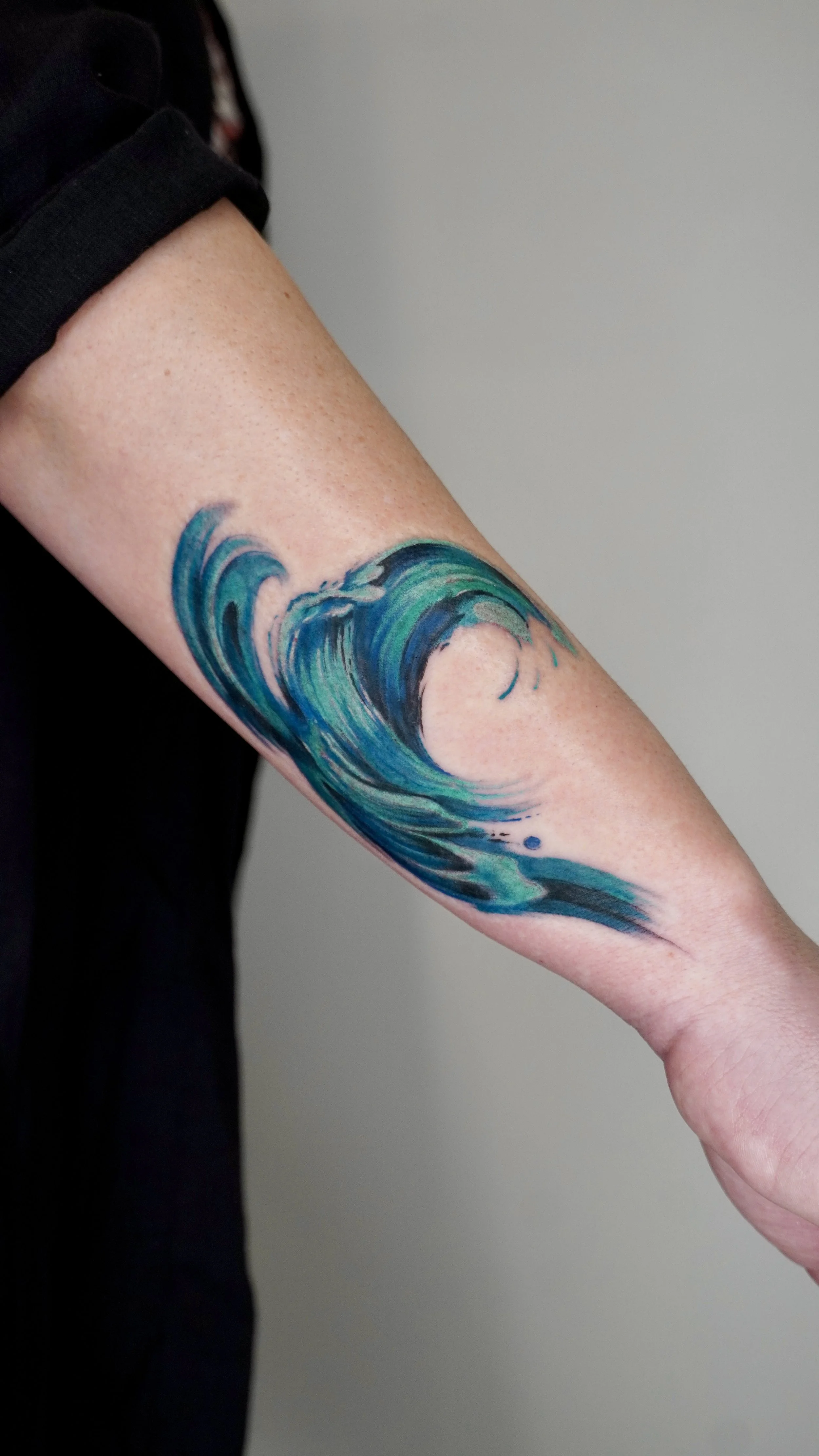 Tattoo of a large ocean wave in shades of blue and green on a person's forearm.