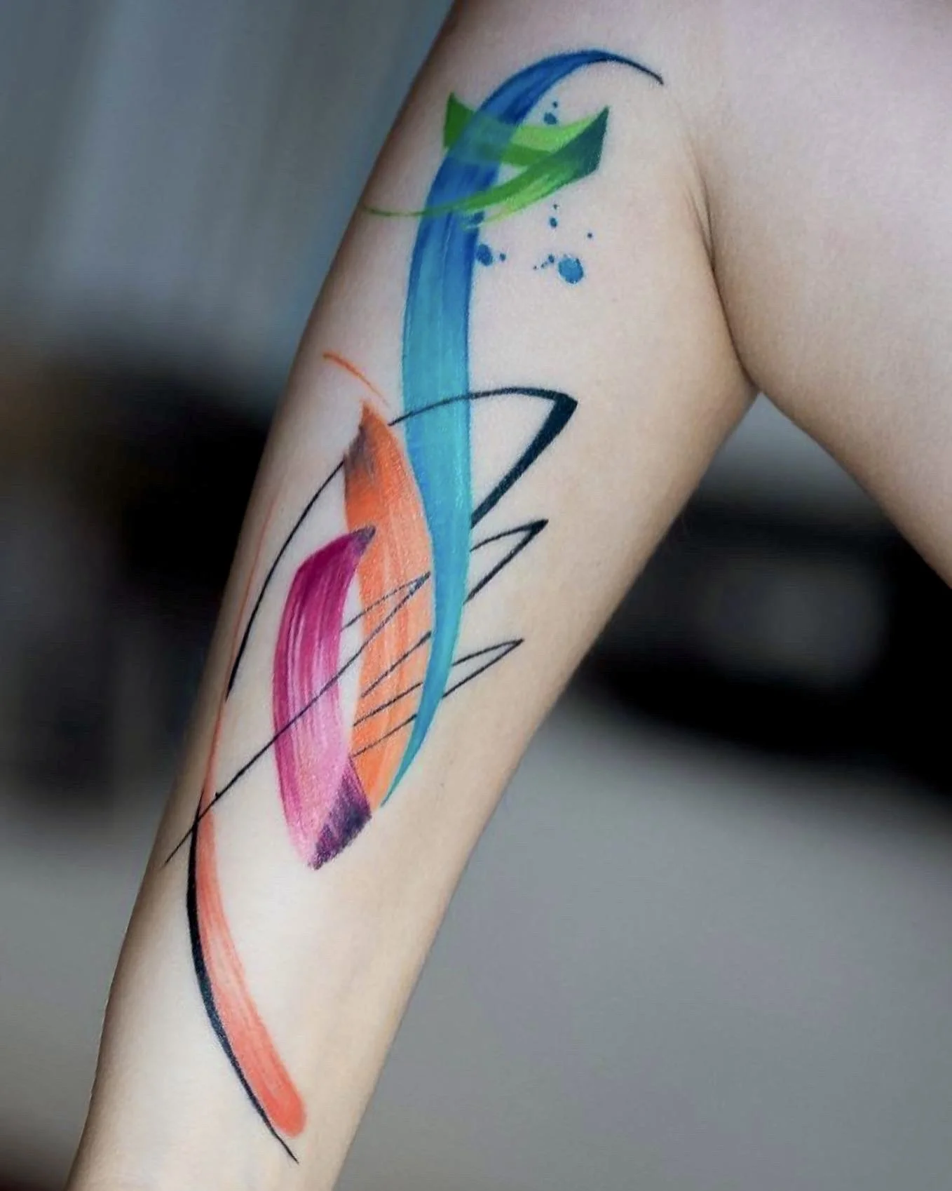 Colorful abstract tattoo on person's forearm, with various brush strokes and splashes of green, blue, orange, pink, and black ink.