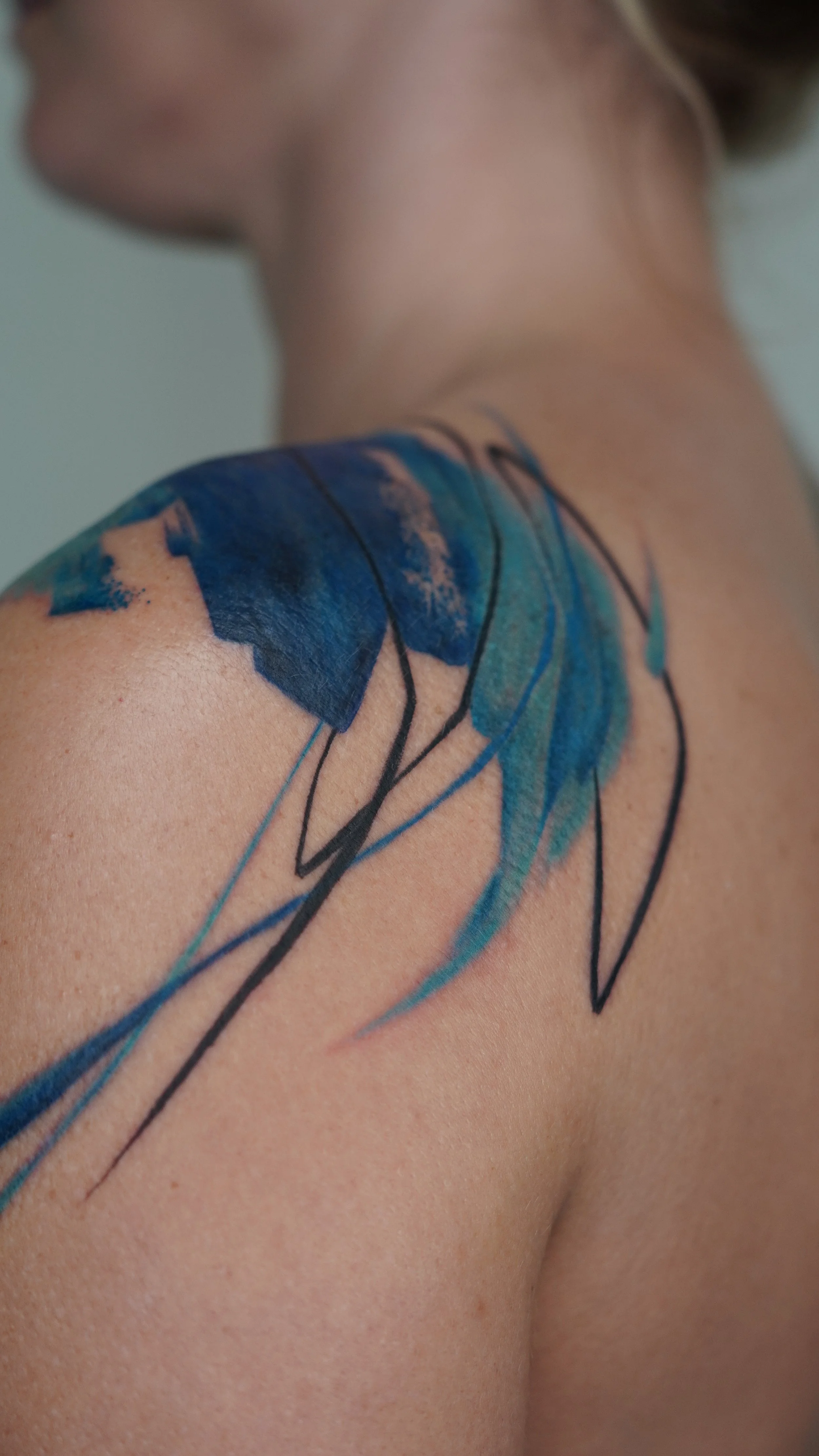 Close-up of a person's shoulder with a watercolor-style tattoo of blue and green leaves and black outlines.