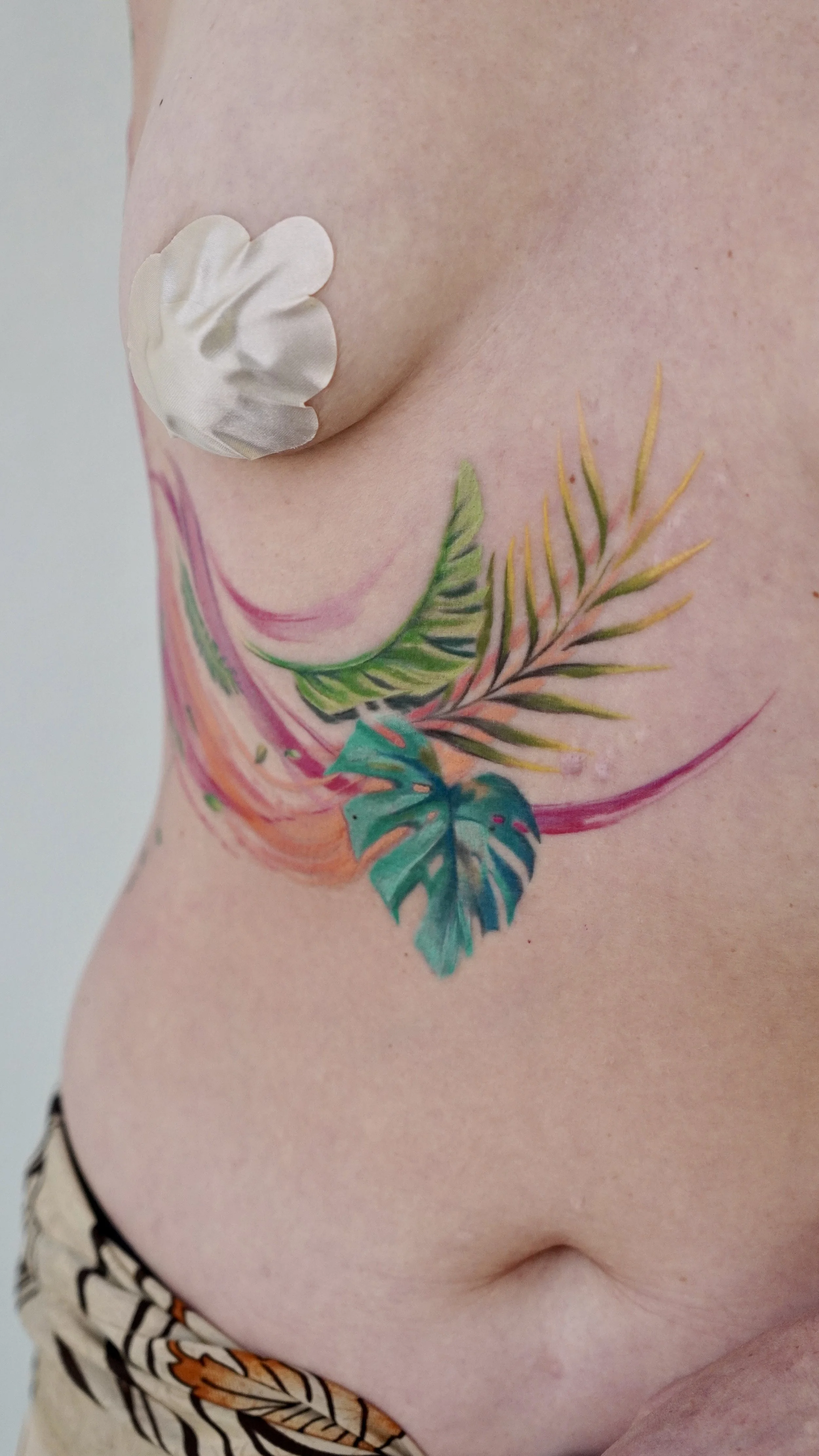 A close-up of a person's side showing a colorful watercolor style tattoo of tropical leaves on their ribs, with a small portion of floral patterned clothing visible at the bottom and a satin patch on their chest.