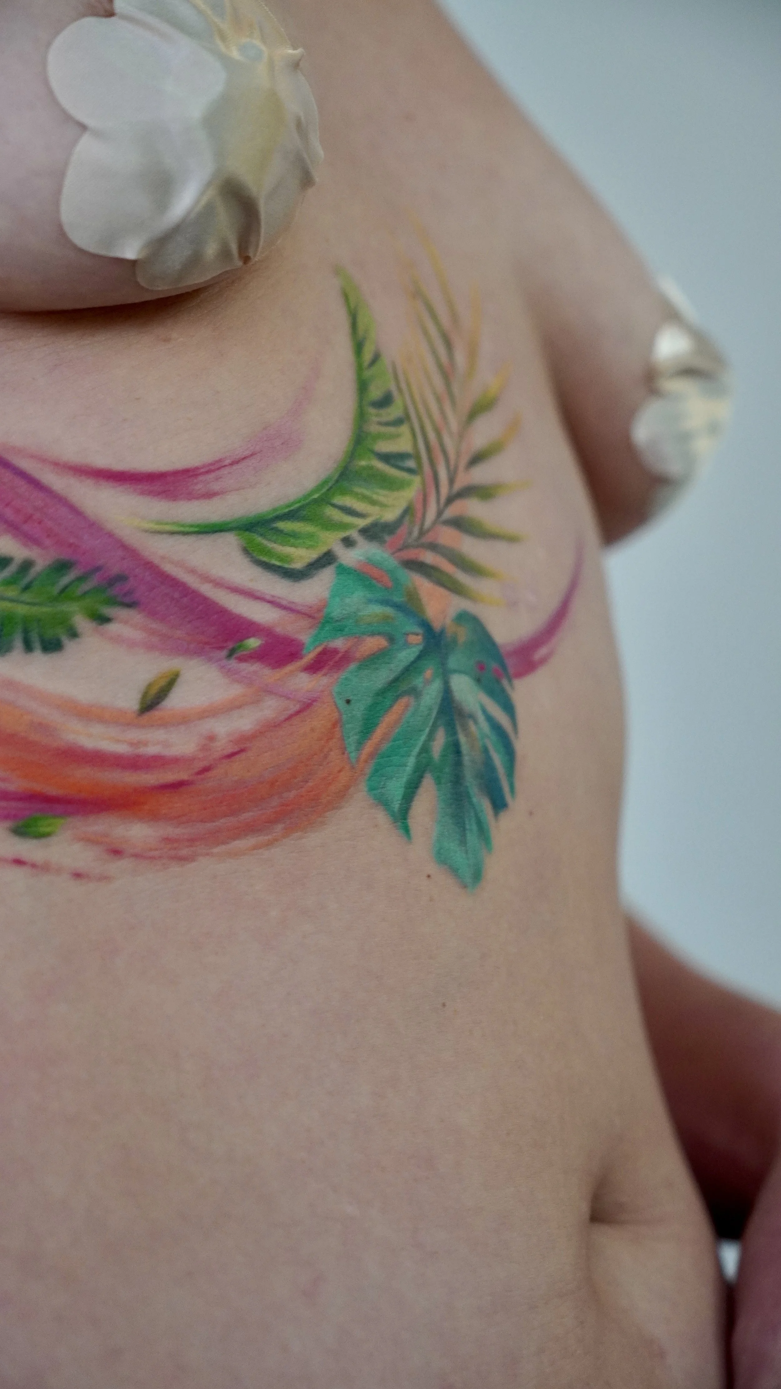Close-up of colorful watercolor-style tropical leaves and swirls tattoo on a person's shoulder and chest, with seashells attached to the shoulder.
