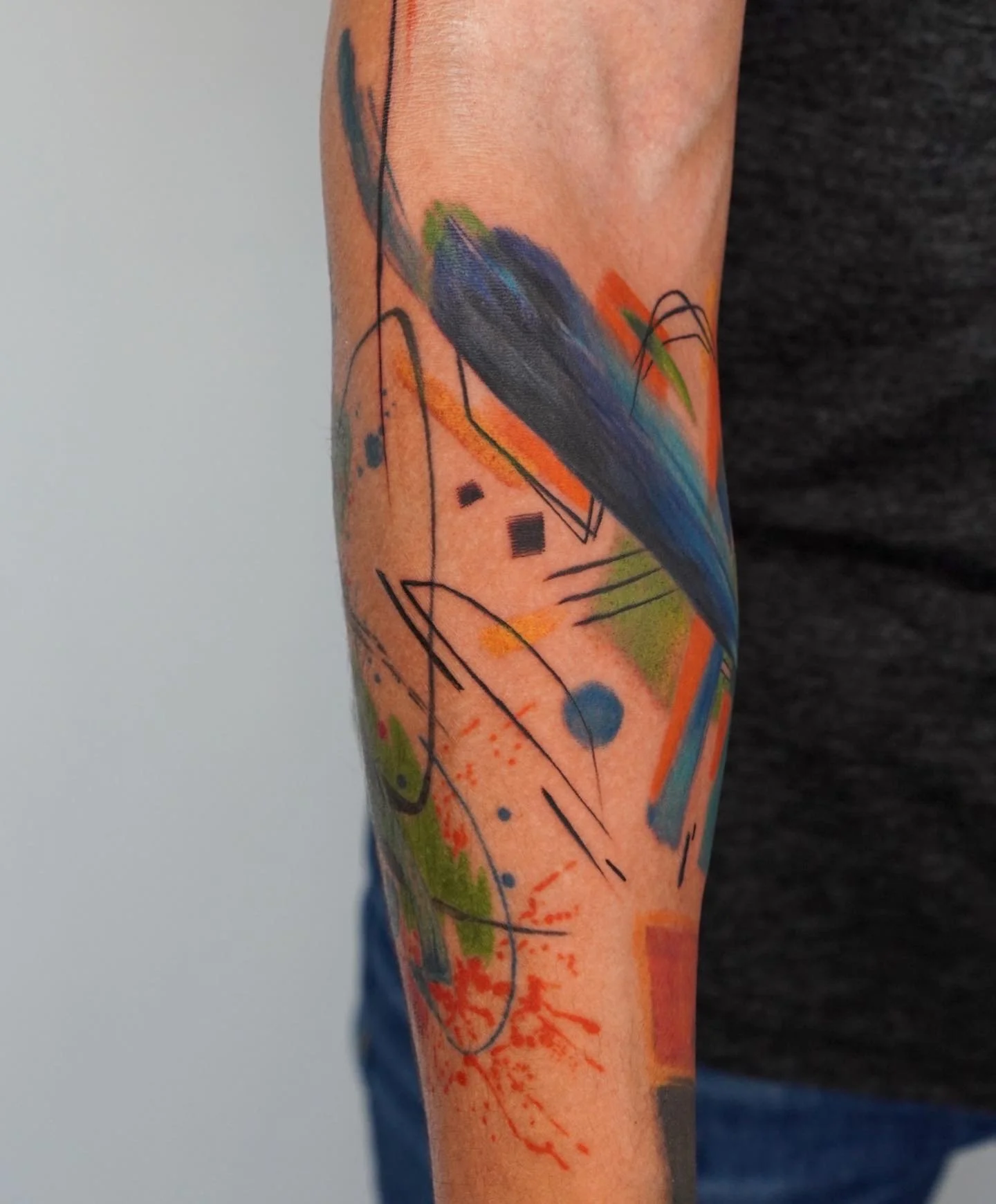 Close-up of a colorful abstract tattoo on a person's arm, featuring splashes of blue, green, orange, black lines, and geometric shapes.