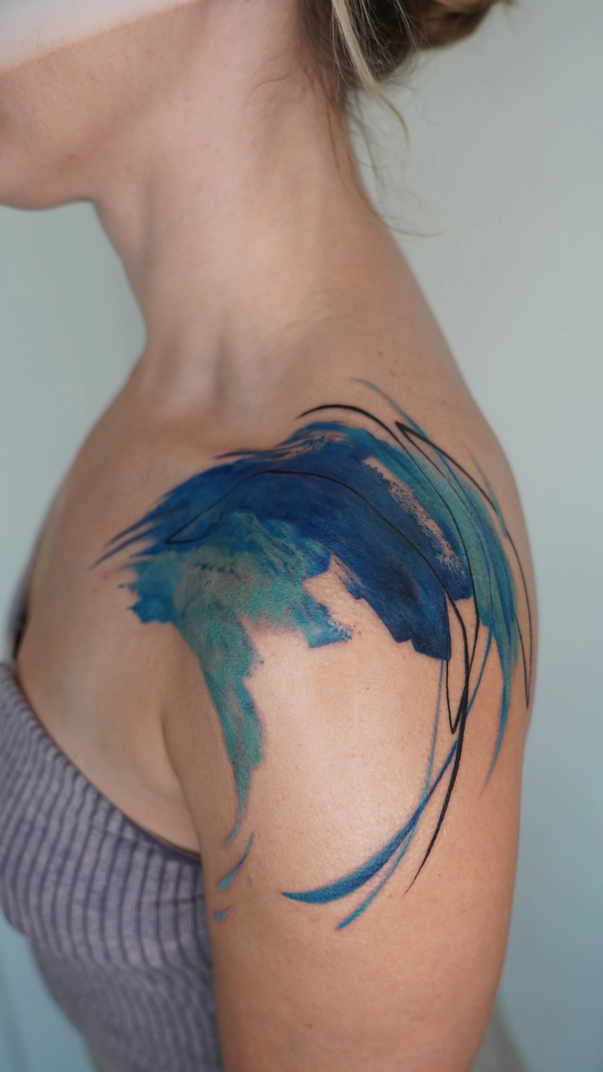 A shoulder tattoo of a stylized, abstract wave painted in shades of blue and green with black accents.