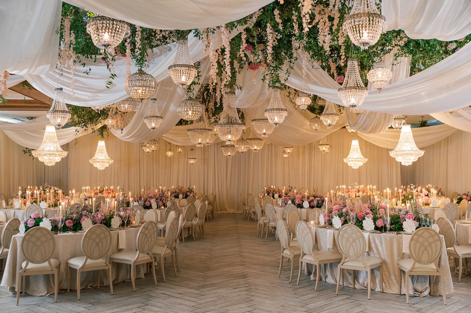 Elegant banquet hall decorated with multiple crystal chandeliers, white drapery, and hanging pink and white flowers with greenery, set for a wedding or formal event.