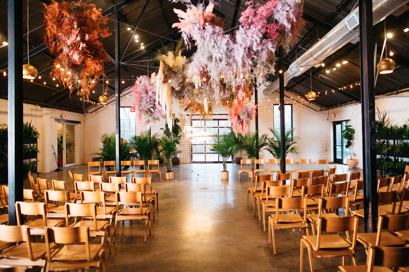 Indoor event space decorated with hanging pink, orange, and white floral arrangements, string lights, and potted plants, with wooden chairs arranged in rows facing a small stage area.