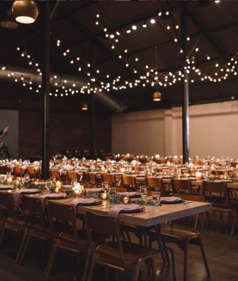 A large banquet hall decorated with string lights, long tables set for a formal event with candles, greenery, plates, and glasses.