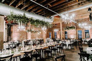 Event venue with long tables, chairs, string lights, and hanging plants in a brick-walled space with wooden ceiling rafters.