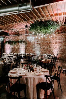 Event space with round tables, white tablecloths, and chairs in a brick-walled room decorated with hanging greenery and string lights.