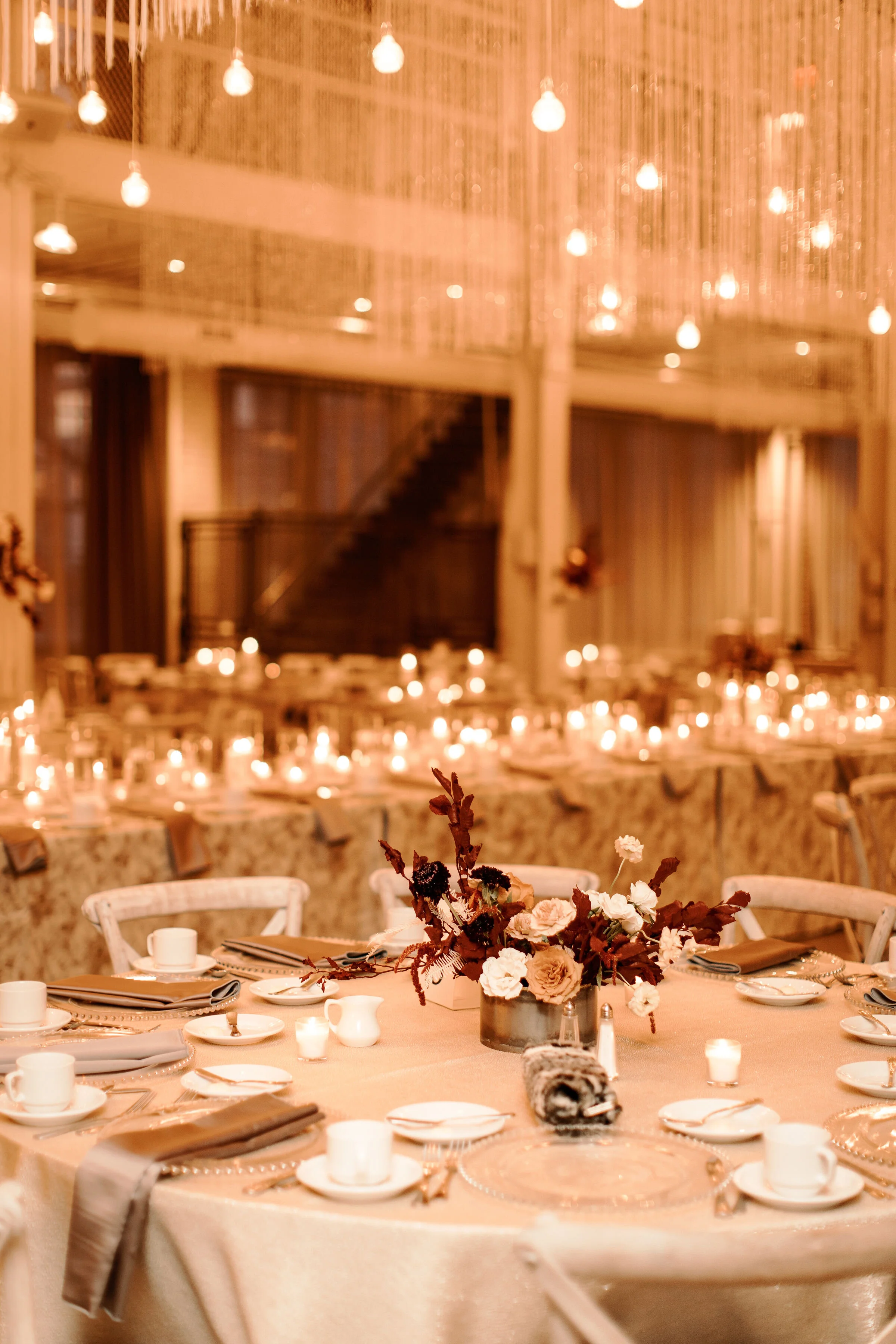 Elegant banquet table decorated with a floral centerpiece, plates, cups, and cutlery, with candles and string lights creating a warm ambiance.