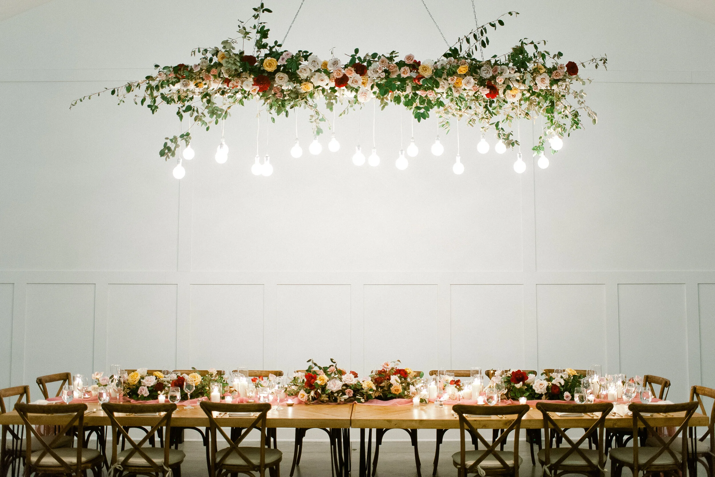 Long wooden dining table with floral centerpiece, candles, and glassware, set for a celebration. Overhead light fixture with hanging light bulbs and floral decoration.