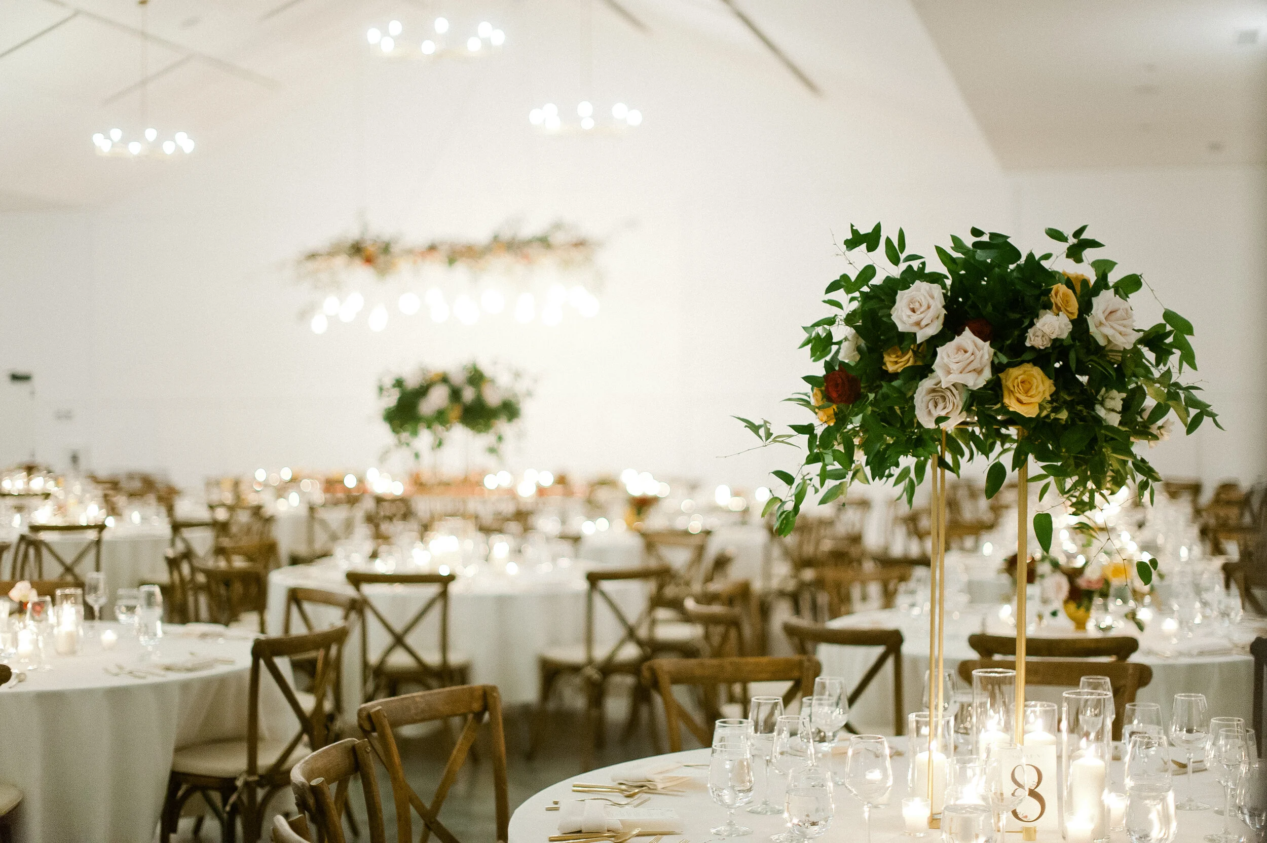 Elegant event space decorated with round tables covered in white tablecloths, each set with glassware, candles, and floral centerpieces, featuring a tall floral arrangement with white, yellow, and dark red roses and green foliage in the foreground.