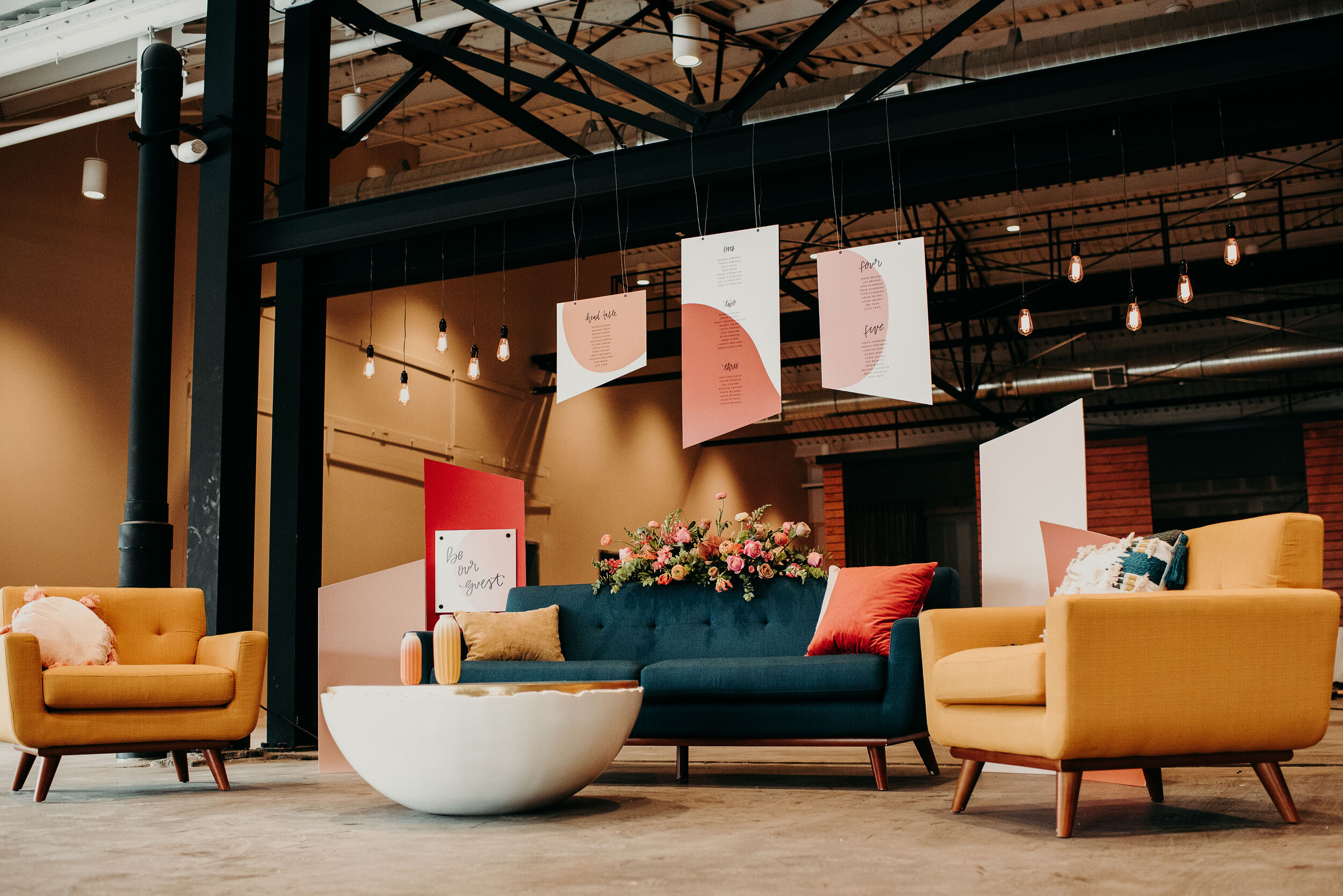 Indoor modern lounge area with yellow and dark teal sofas, a white coffee table, a flower arrangement, and hanging paper decorations with text, illuminated by string lights.
