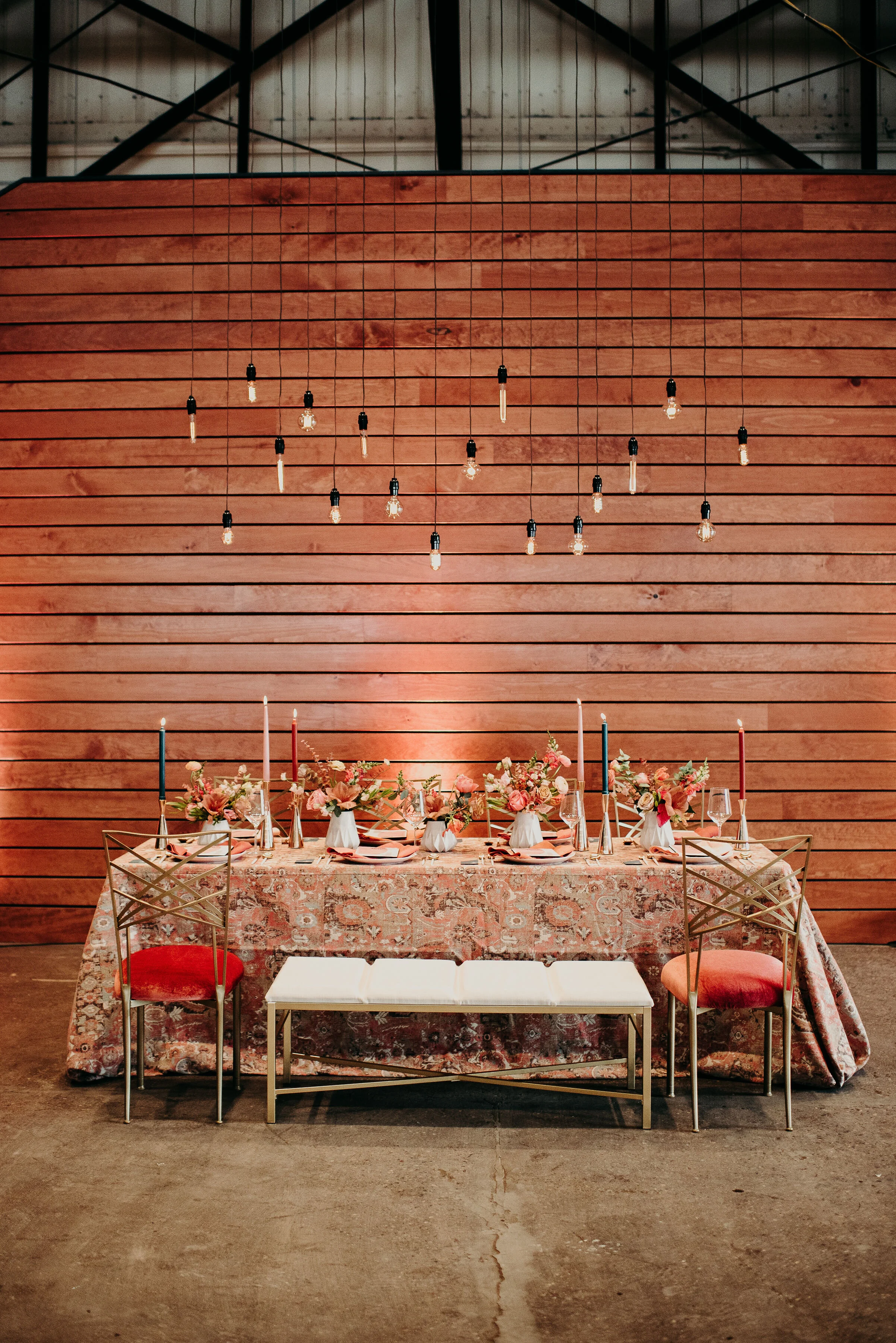 A decorated dining table set for a special occasion with pastel pink and red flowers, tall candles in pink and blue, and glassware, against a wooden backdrop with hanging Edison bulbs.