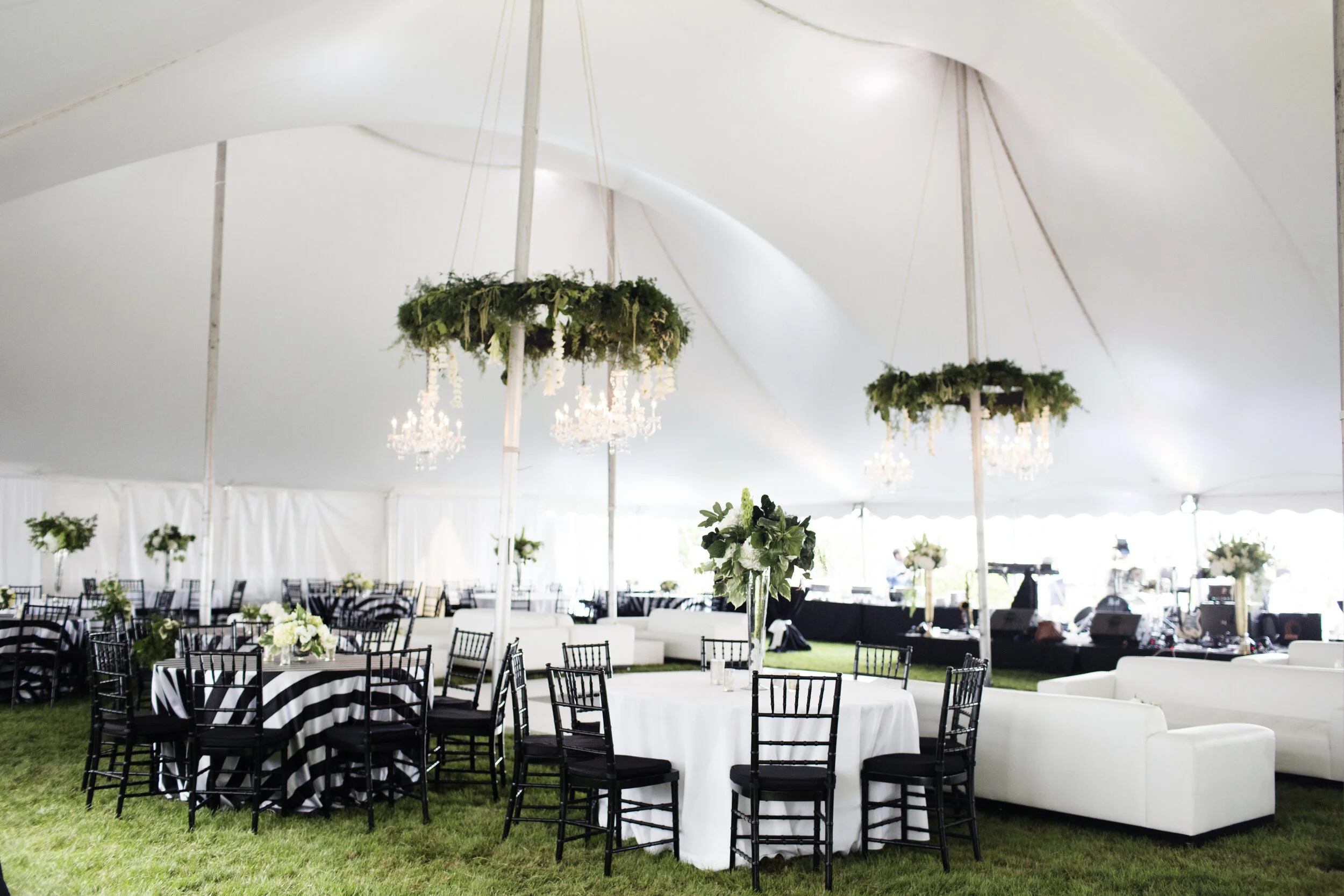 Elegant event tent with round tables covered in black and white striped tablecloths, decorated with floral centerpieces; white couches and black chairs arranged on green grass; chandeliers hanging from the ceiling, and floral chandeliers hanging from