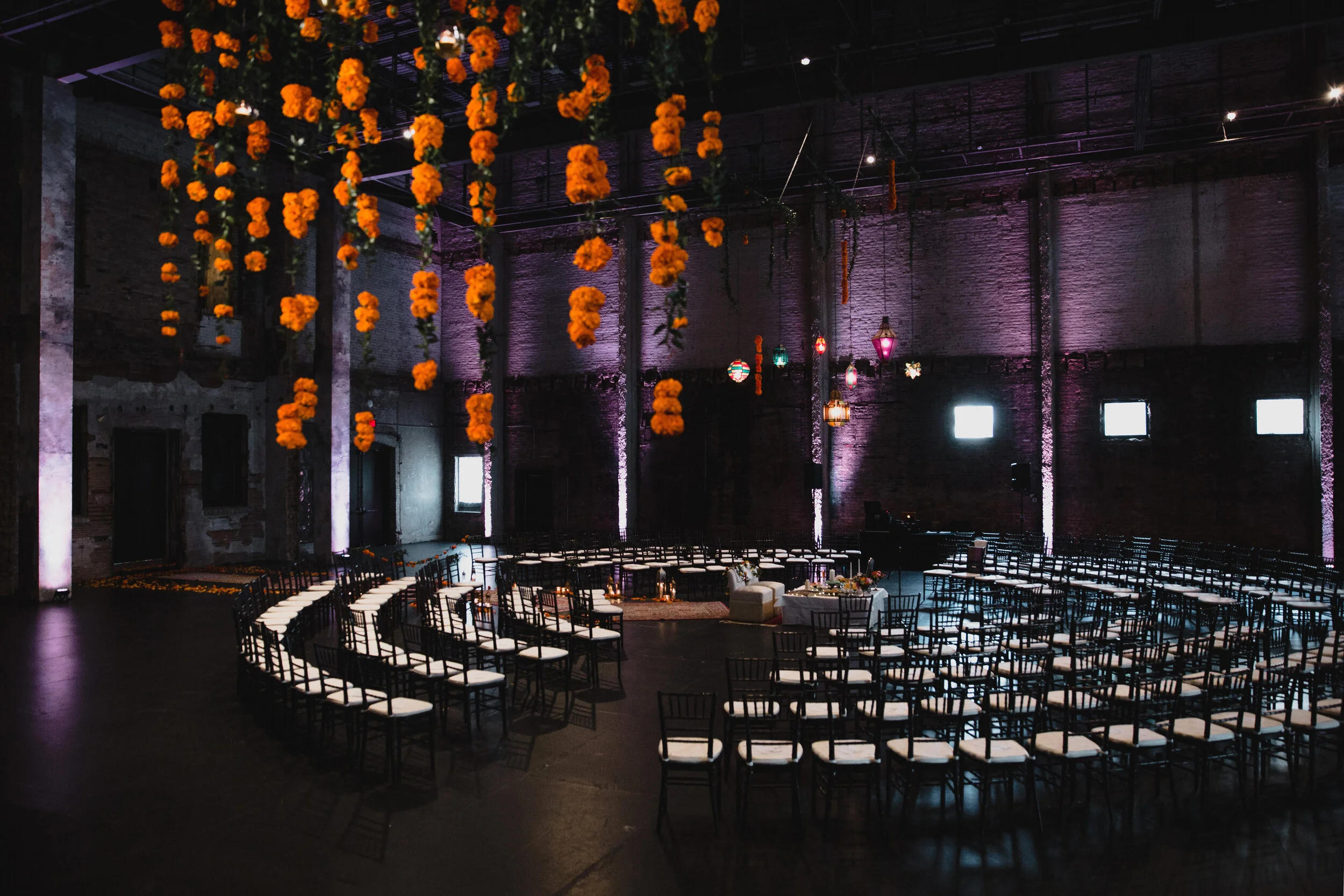 A large indoor event space decorated with hanging orange marigold garlands and colorful lanterns, with rows of black chairs with white cushions arranged in semi-circles, and a lounge area with a sofa and table, ready for a celebration or wedding.