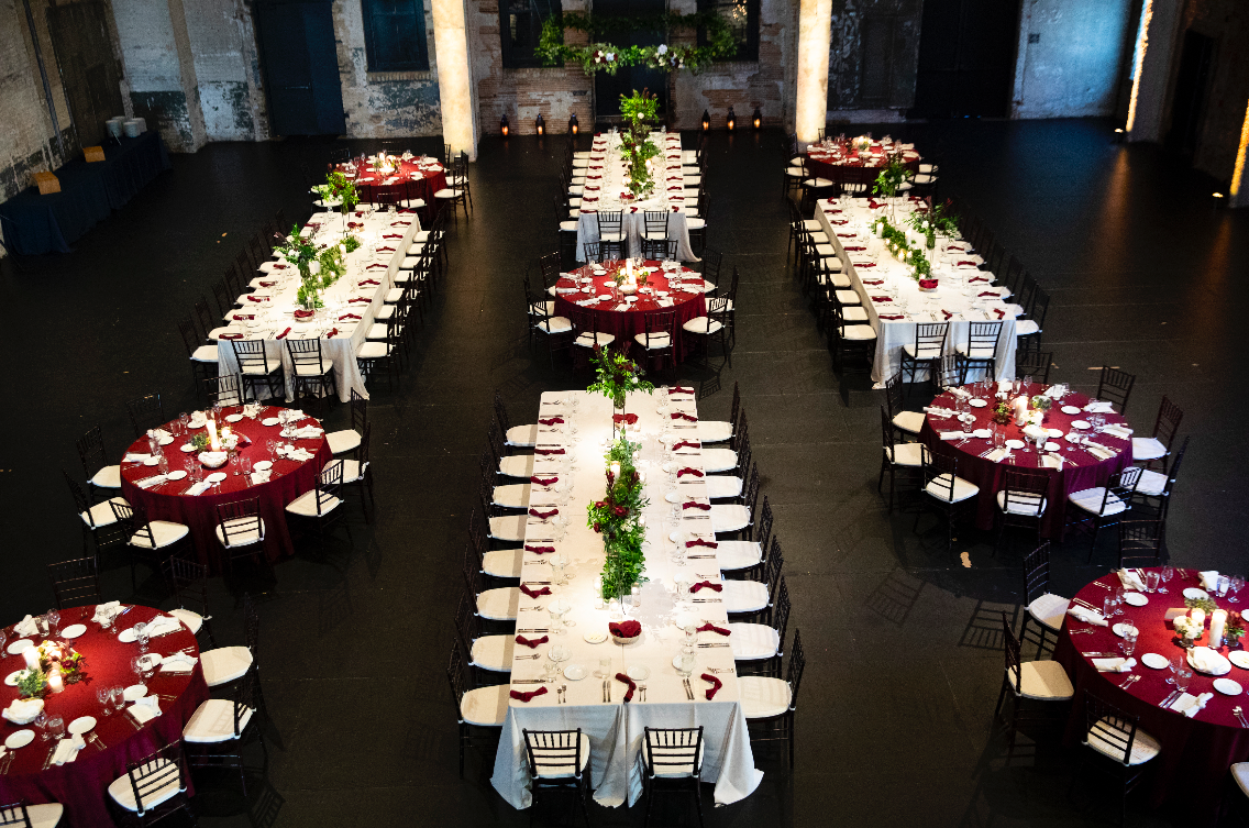 Elegant banquet setup with long and round tables decorated with white tablecloths, burgundy runners, and green floral centerpieces inside a rustic venue.