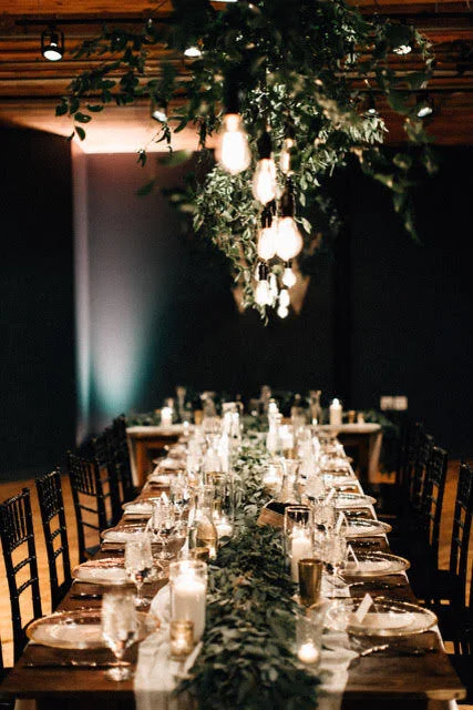 Elegant indoor dining setup with a long wooden table, candles, greenery, and hanging lights for a wedding or event.