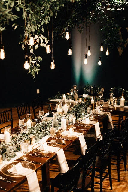 Elegant party table setup with lit hanging bulbs, greenery centerpiece, wine glasses, plates, and napkins on a wooden table in a dimly lit room.