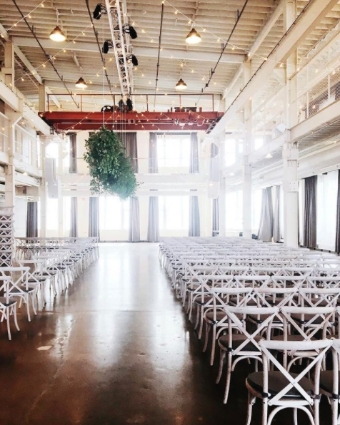 Empty event space or conference room with rows of white chairs, large windows with curtains, and a large hanging plant with industrial ceiling lighting.