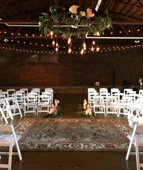 Indoor wedding ceremony setup with white chairs arranged on either side of an aisle, a floral chandelier hanging from the ceiling, and string lights overhead. There is a decorative rug on the floor, and candles and greenery at the front of the aisle.