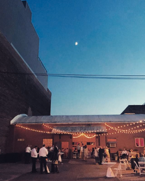 Evening outdoor event with guests socializing under string lights in an urban setting, with buildings and a clear night sky with visible moon.