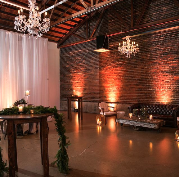 Elegant indoor event space with exposed brick wall, chandeliers, string lights, and cozy seating including armchairs and a sofa, decorated with candles and greenery.