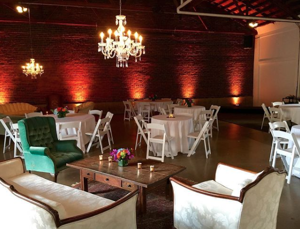 Elegant event space with brick walls, chandeliers, and round tables with white tablecloths and floral centerpieces.