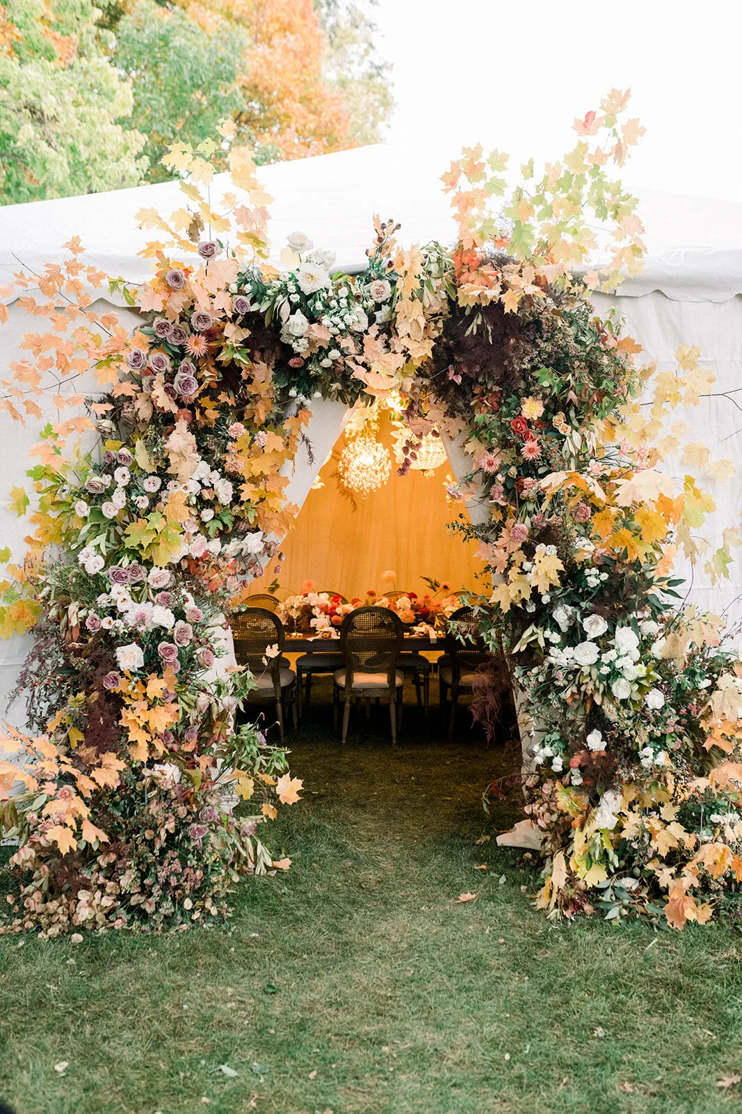 Decorated wedding arch with flowers and autumn leaves, overlooking a table set for a celebration inside a white tent, with trees showing fall colors in the background.